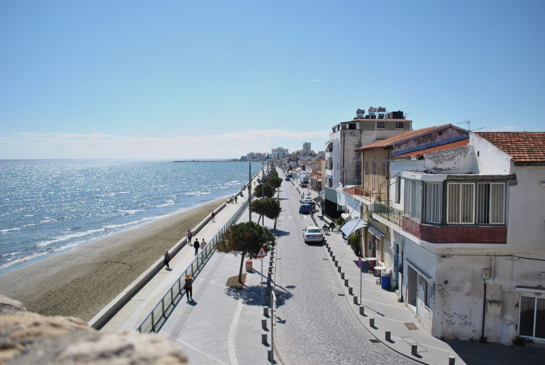 A seafront promenade in a Cyprus coastal city seen from above, with a long sand beach and calm blue sea on one side and old low houses lining the road on the other