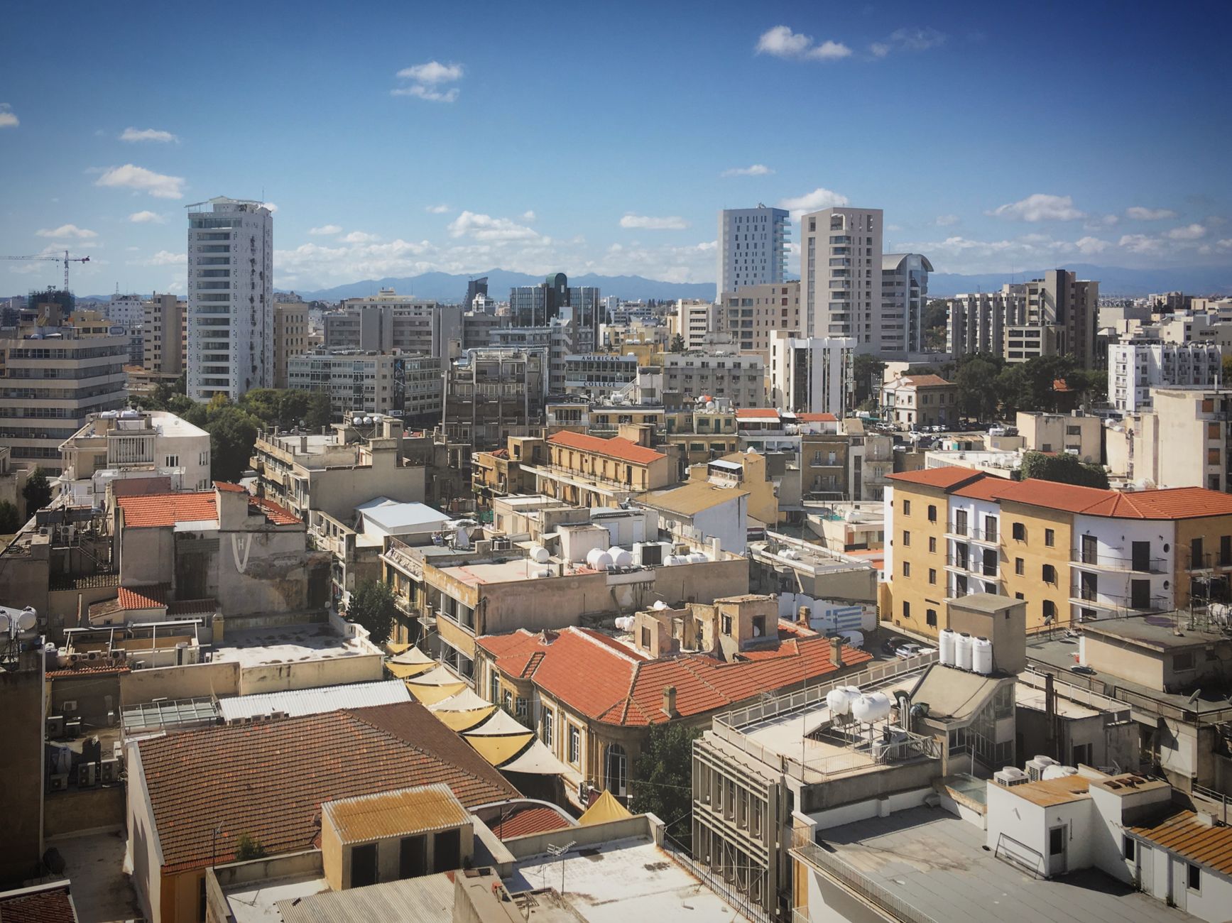 A Cypriot city seen from above, with white apartment towers and a construction crane rising behind older red-tiled rooftops and a mountain range on the horizon