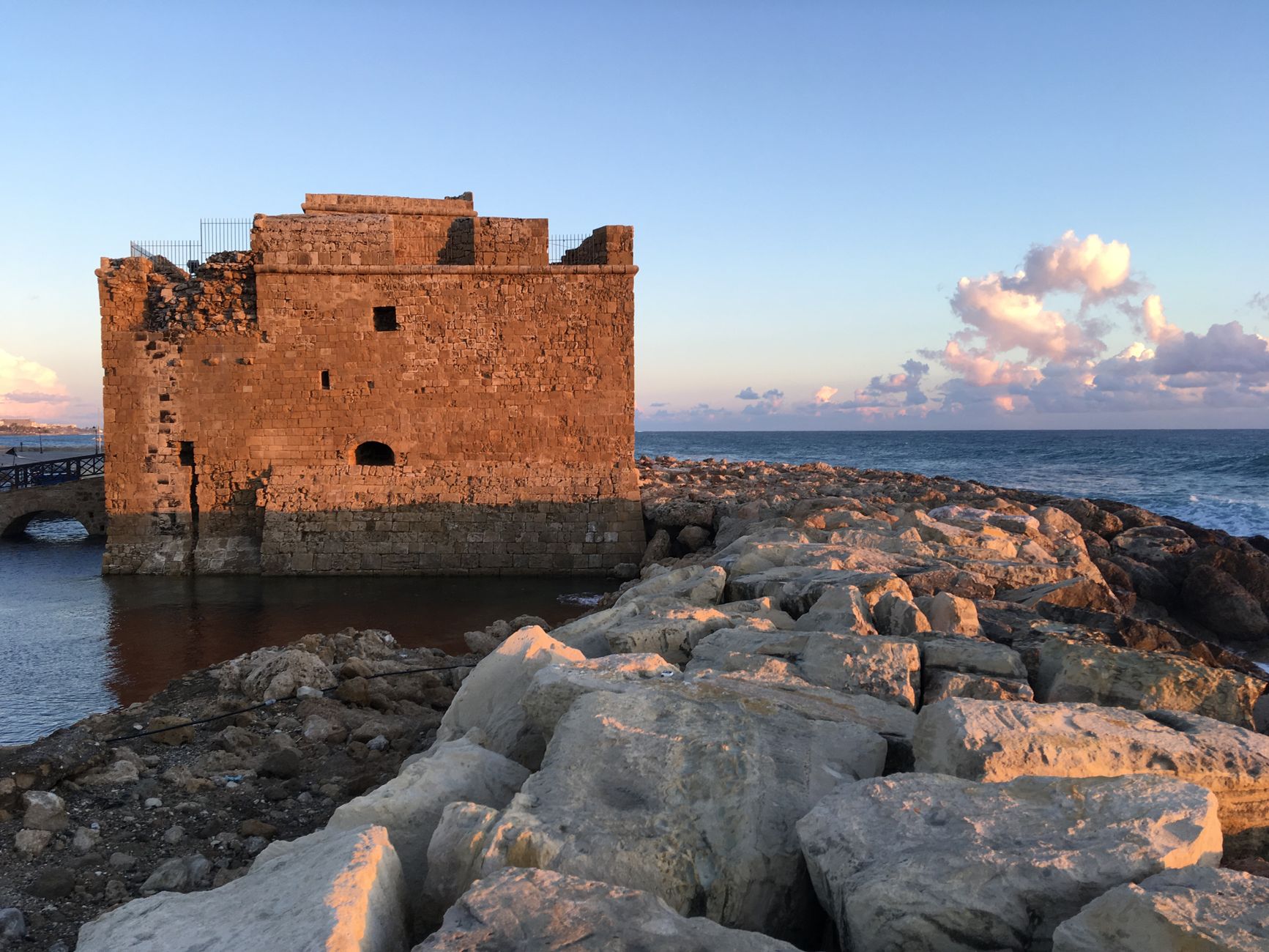 A weathered stone medieval castle standing on the Mediterranean shoreline at sunset, with a pale limestone breakwater in front of it and open sea behind