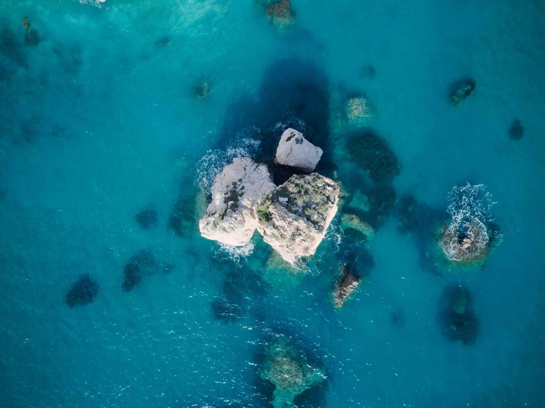 Overhead aerial view of a pale limestone outcrop breaking the surface of clear turquoise water, with submerged reefs and their shadows spreading across the seabed around it