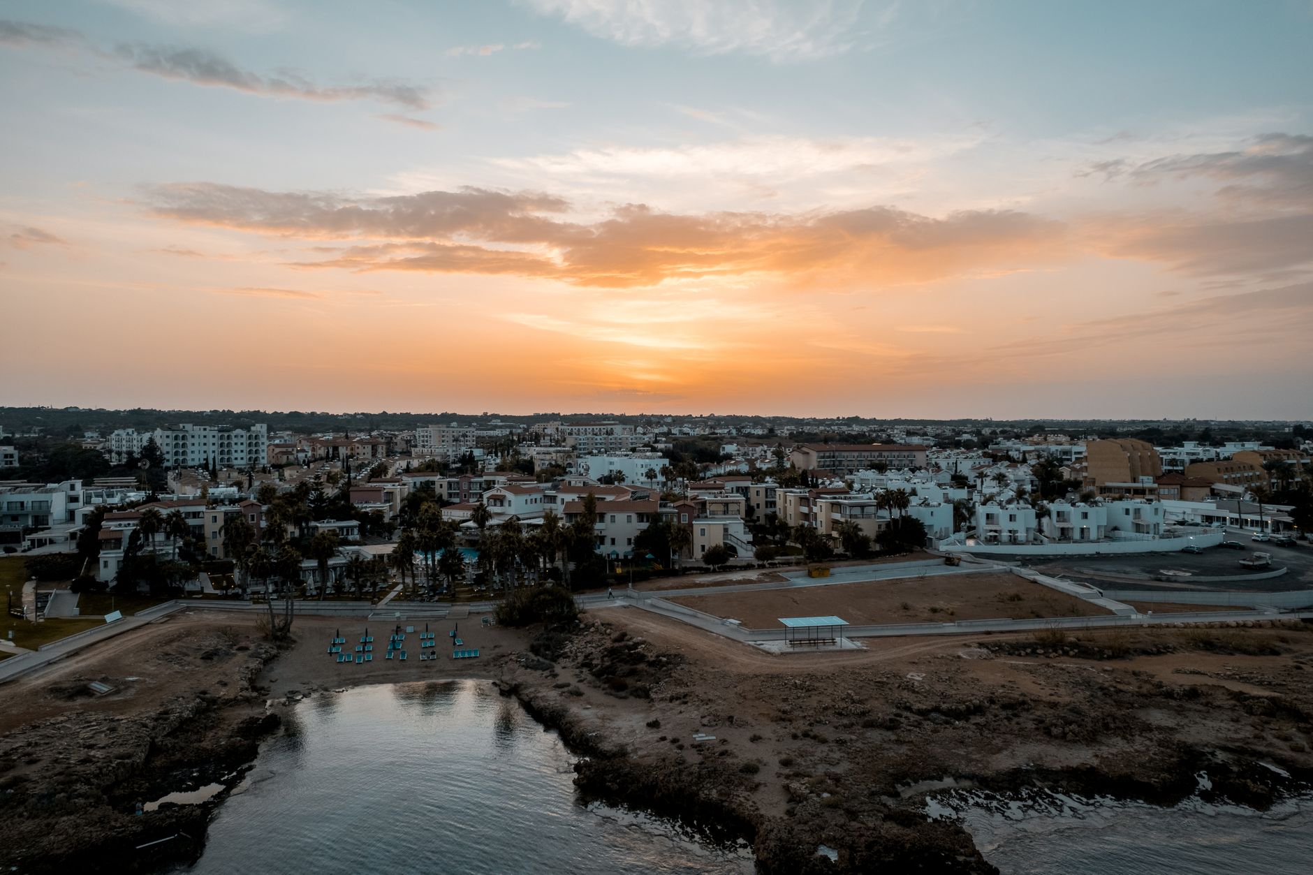 Aerial view at sunset over a low-rise Cyprus coastal town of white apartment blocks and terracotta roofs, with palm trees and a small rocky swimming cove in the foreground