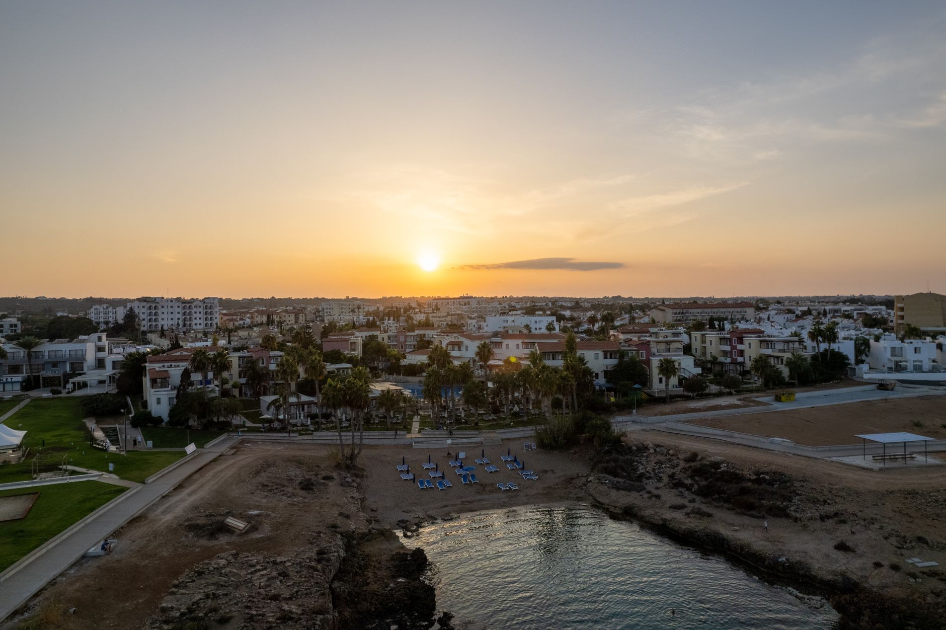 Sunset over a Cyprus seaside town photographed from above, terracotta roofs and palm-lined gardens above a small rocky inlet
