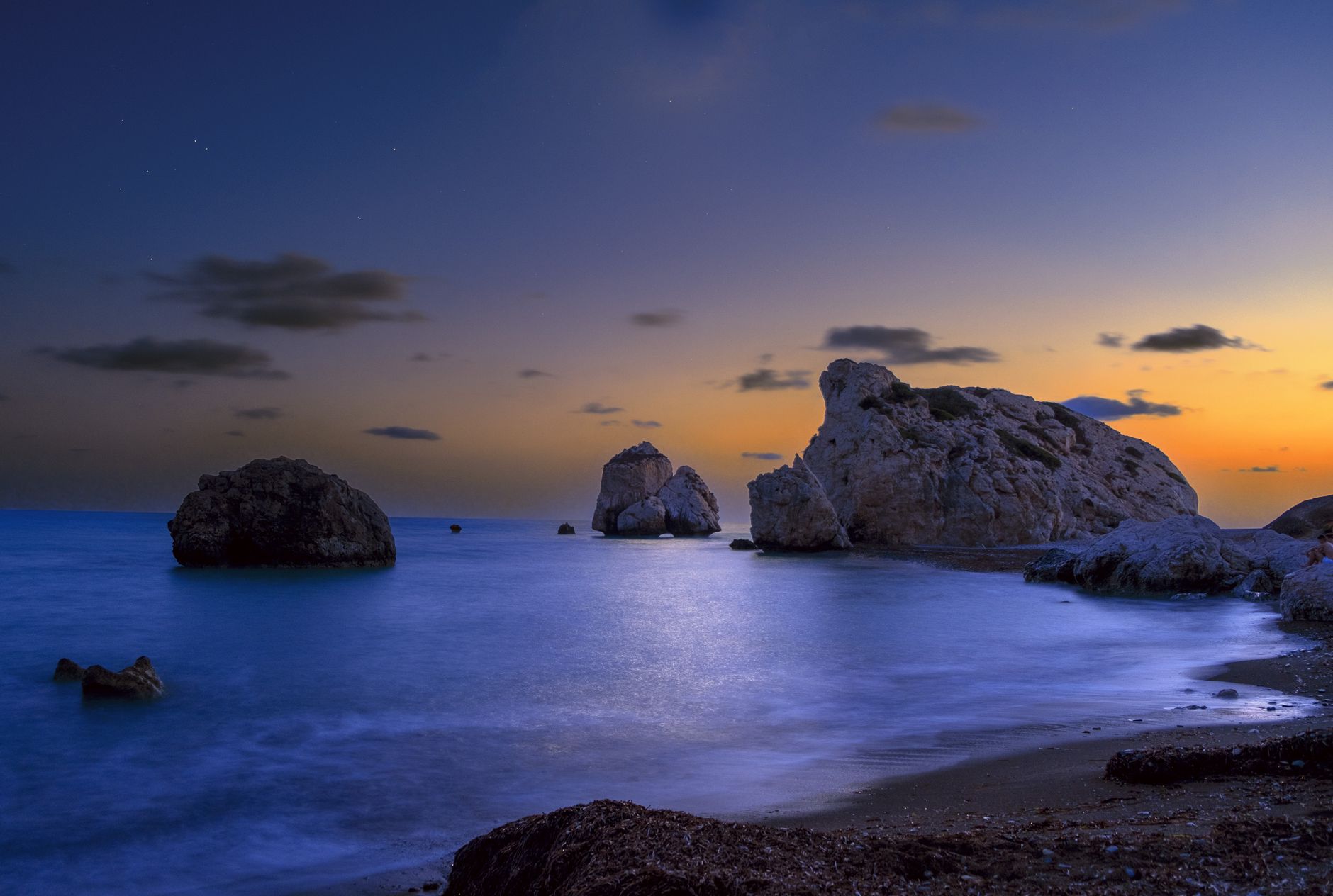 Weathered sea stacks standing in calm shallow water at dusk, an orange band of afterglow along the horizon and a dark sandy shore curving into the foreground