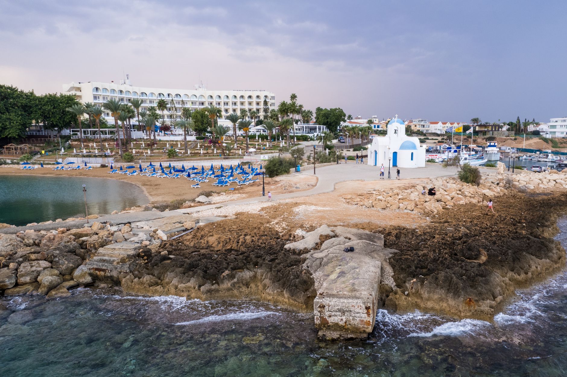 A Mediterranean resort bay from the air at dusk: a long curved white hotel behind palm trees, rows of blue sunbeds on a small sandy beach, and a whitewashed chapel with a blue dome on the rocky point beside moored boats