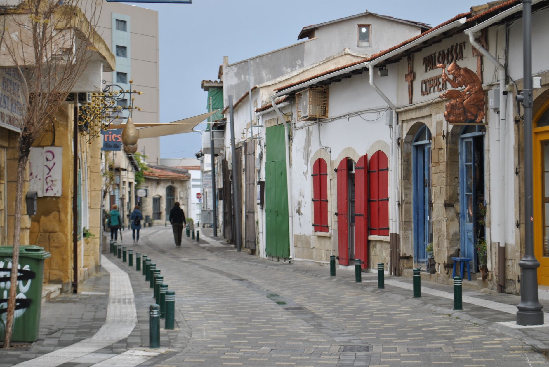 A narrow Mediterranean old-town street of low stone houses with red, green and blue wooden shutters and terracotta roofs, bollards along a paved lane and three people walking away from the camera