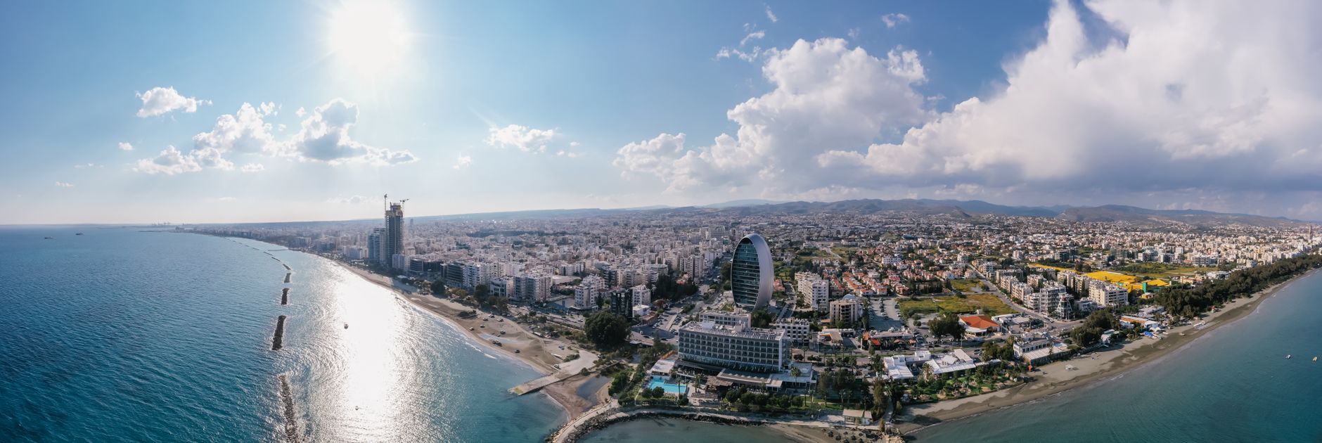 The Limassol seafront from the air, with the elliptical Oval tower and a high-rise under construction along the shore and the Troodos foothills behind