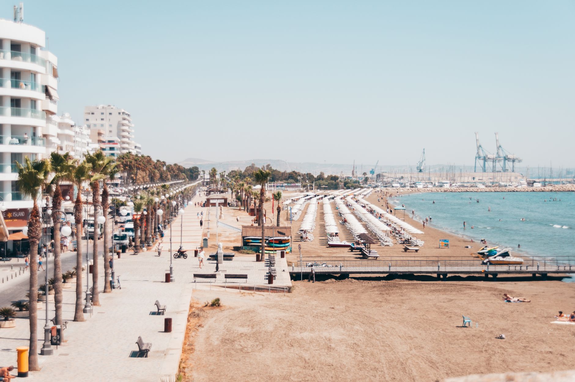 A palm-lined seafront promenade in a Cyprus coastal city, with a marina and port cranes visible in the distance