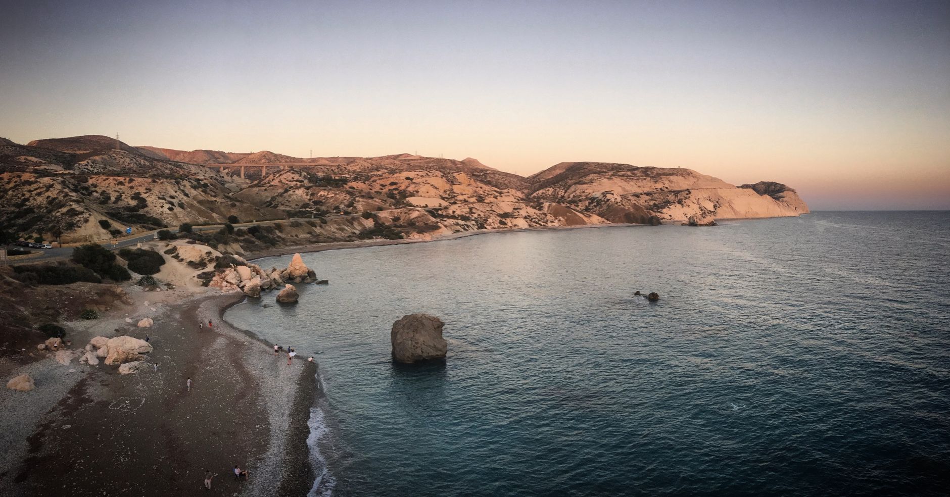 A wide Cyprus bay at dusk, pale limestone headlands and dry scrub hills above a pebble shore, a large sea stack standing alone in calm turquoise water