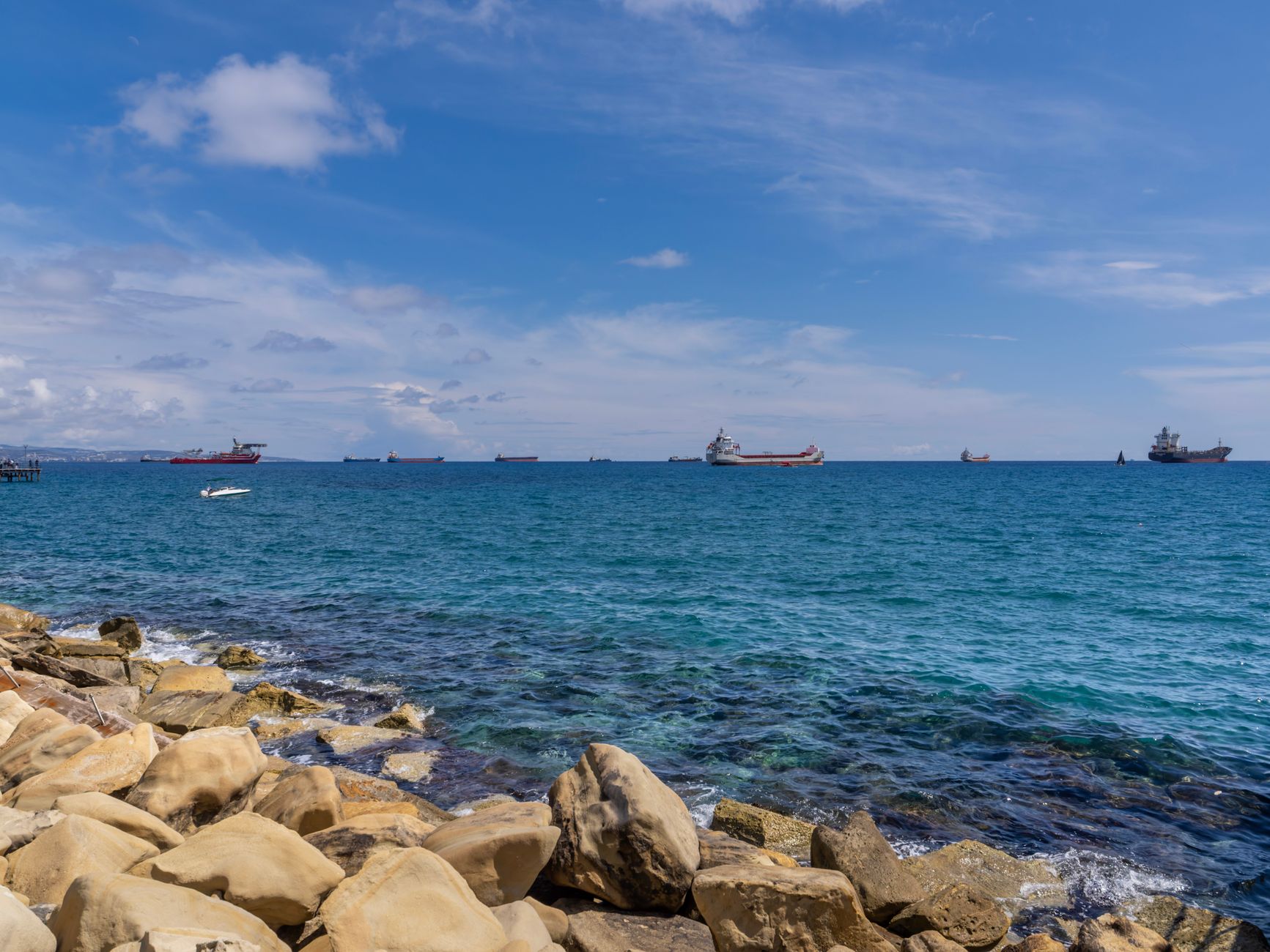 The rocky shoreline of a Cyprus coastal city, with cargo ships and tankers riding at anchor on the horizon and a small white motorboat close to shore