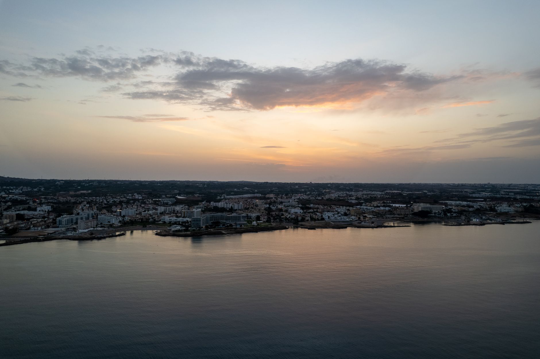 Aerial view at dusk of a low-rise Cyprus coastal town spread along a headland, with hotels and apartment blocks facing a calm sea and an orange sky above the hills inland