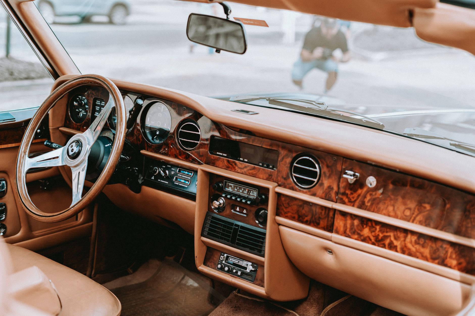 The interior of a vintage saloon in tan leather, with a polished burl-walnut dashboard, round chrome-ringed dials and a slim wooden steering wheel