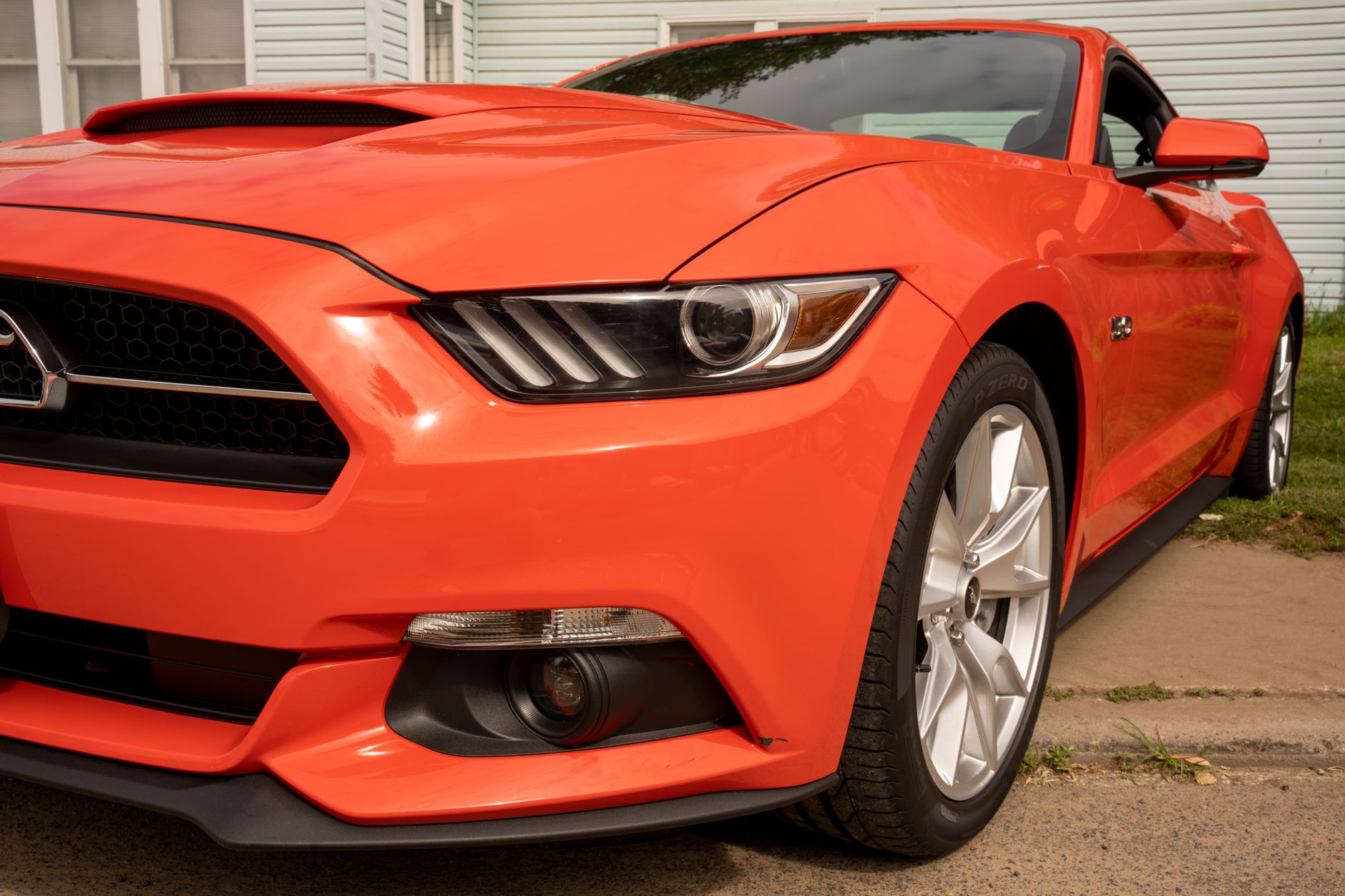 A close view of the front corner of an orange-red sports coupé parked by a kerb, its chrome-rimmed headlight and polished alloy wheel catching the light, with a pale weatherboarded wall behind