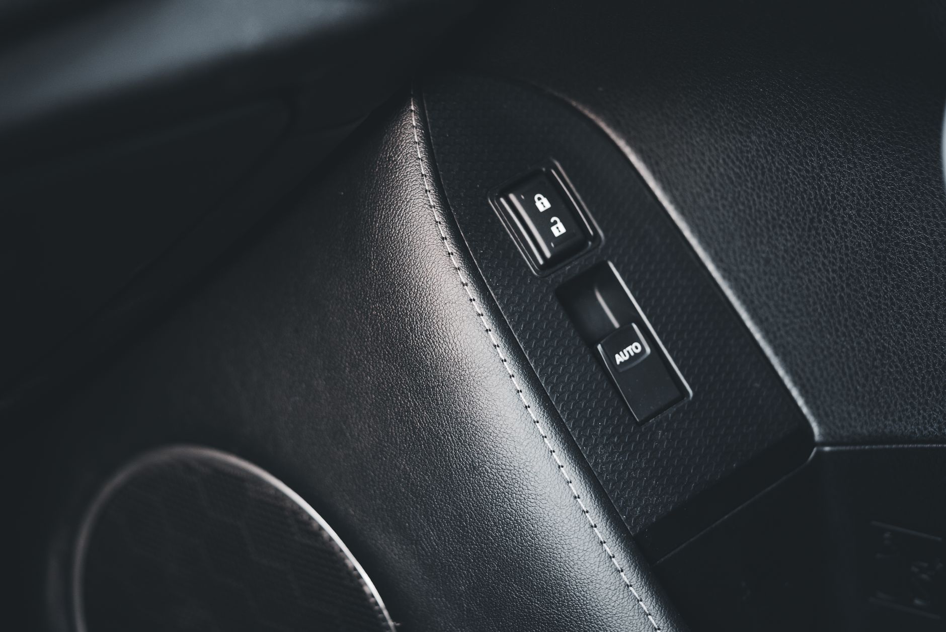 A close-up of a black leather car door panel with pale contrast stitching, a chrome-ringed lock and unlock rocker above a window switch marked AUTO, and the blurred edge of a speaker grille in the foreground