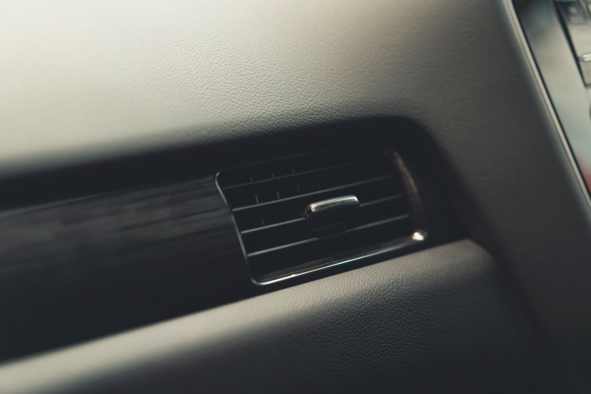 Close-up of a car dashboard air vent set into dark textured leather trim, with a thin chrome slider and a brushed dark panel running alongside it