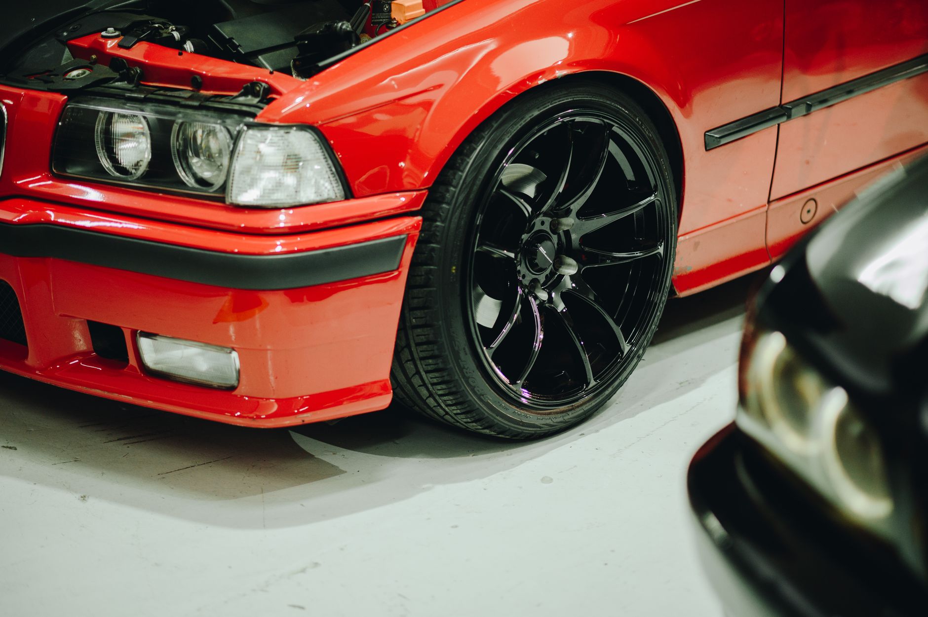 A red coupé with black alloy wheels and its bonnet raised, parked on a pale workshop floor beside a second car