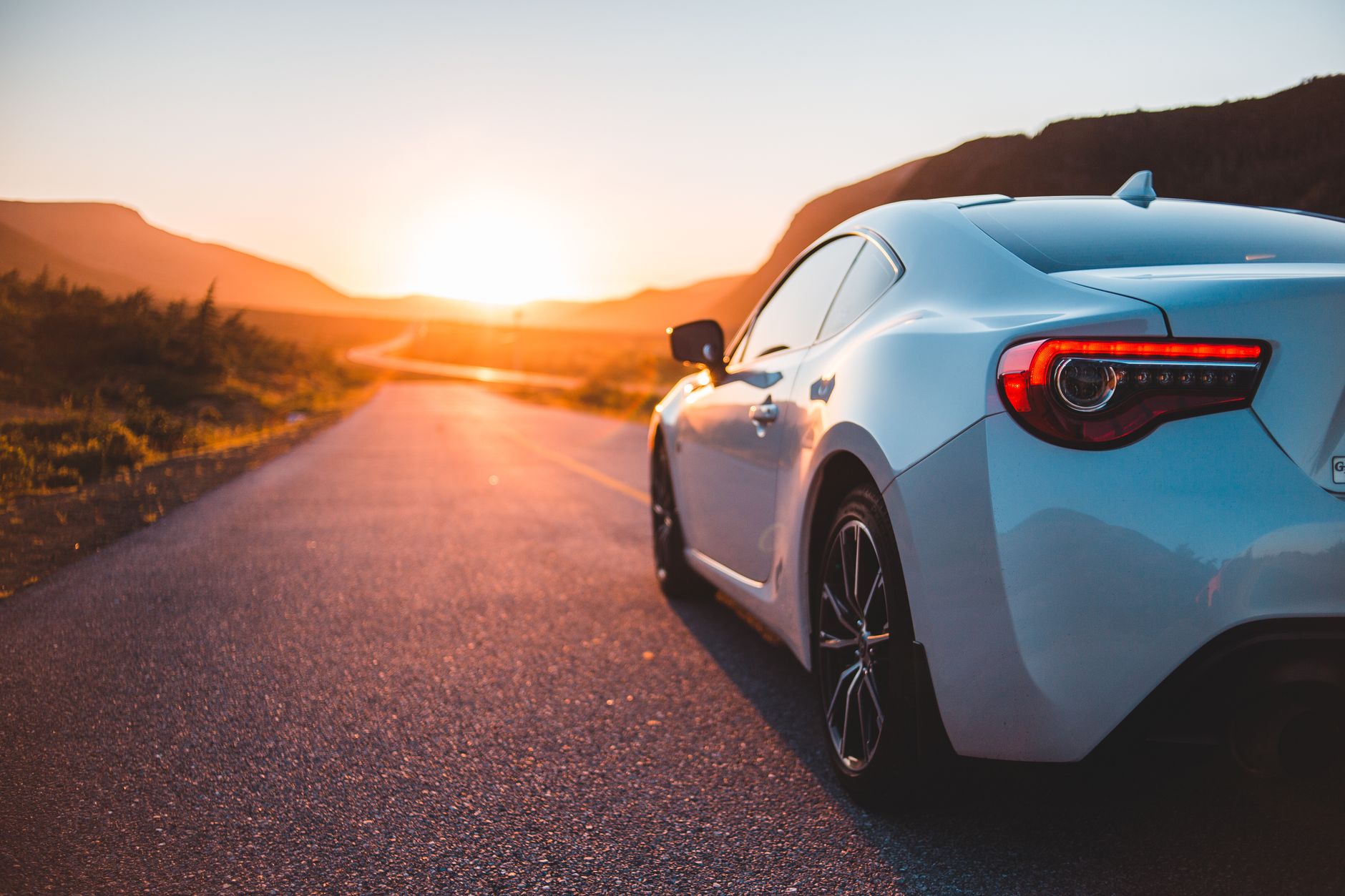 A pale blue sports coupé parked at the roadside on an empty two-lane road at sunset, low hills behind it and the sun on the horizon ahead
