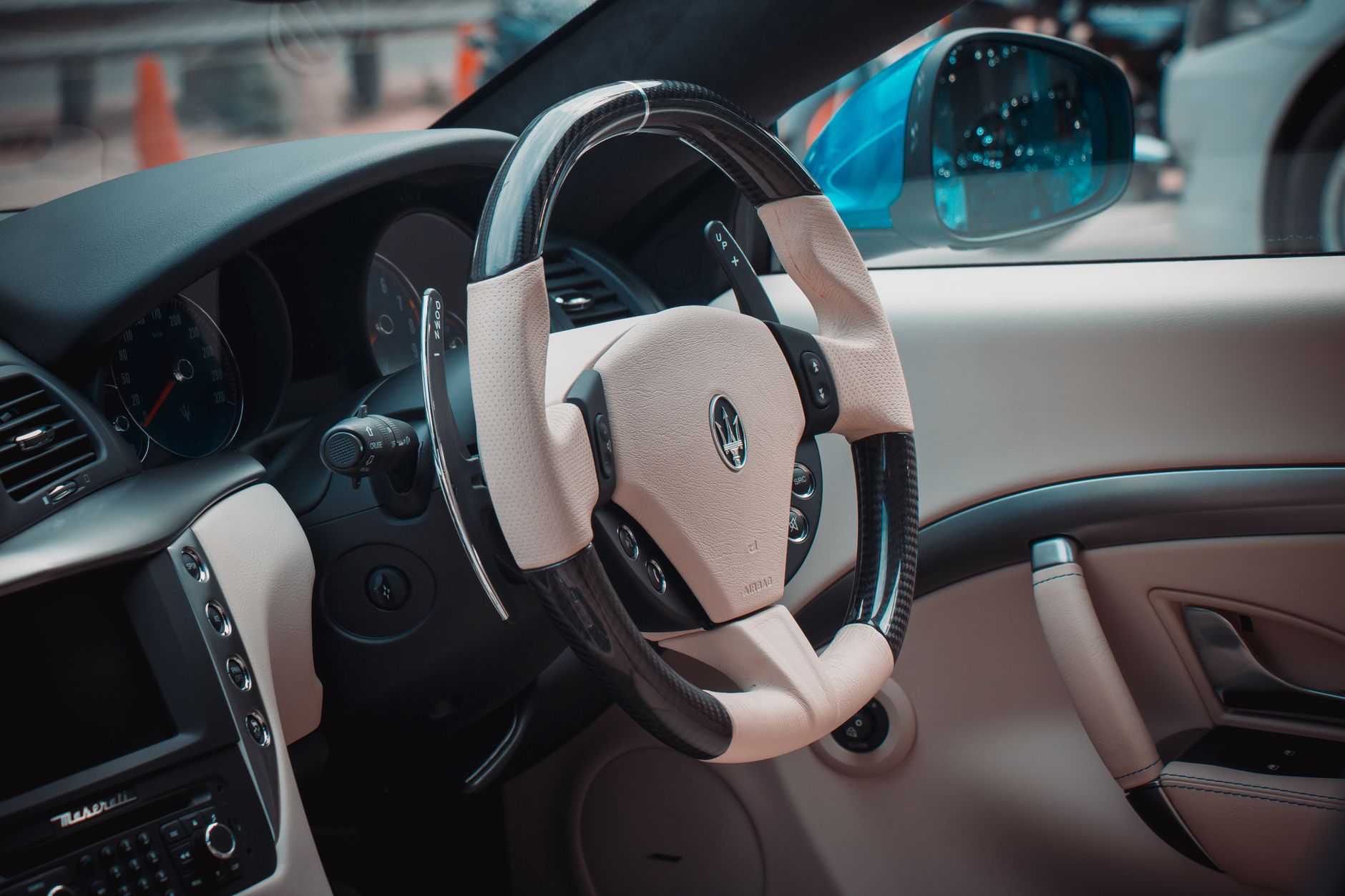Interior of a sports saloon seen from the passenger seat: a cream leather dashboard and door card, a carbon-rimmed steering wheel with paddle shifters, and the instrument dials beyond it