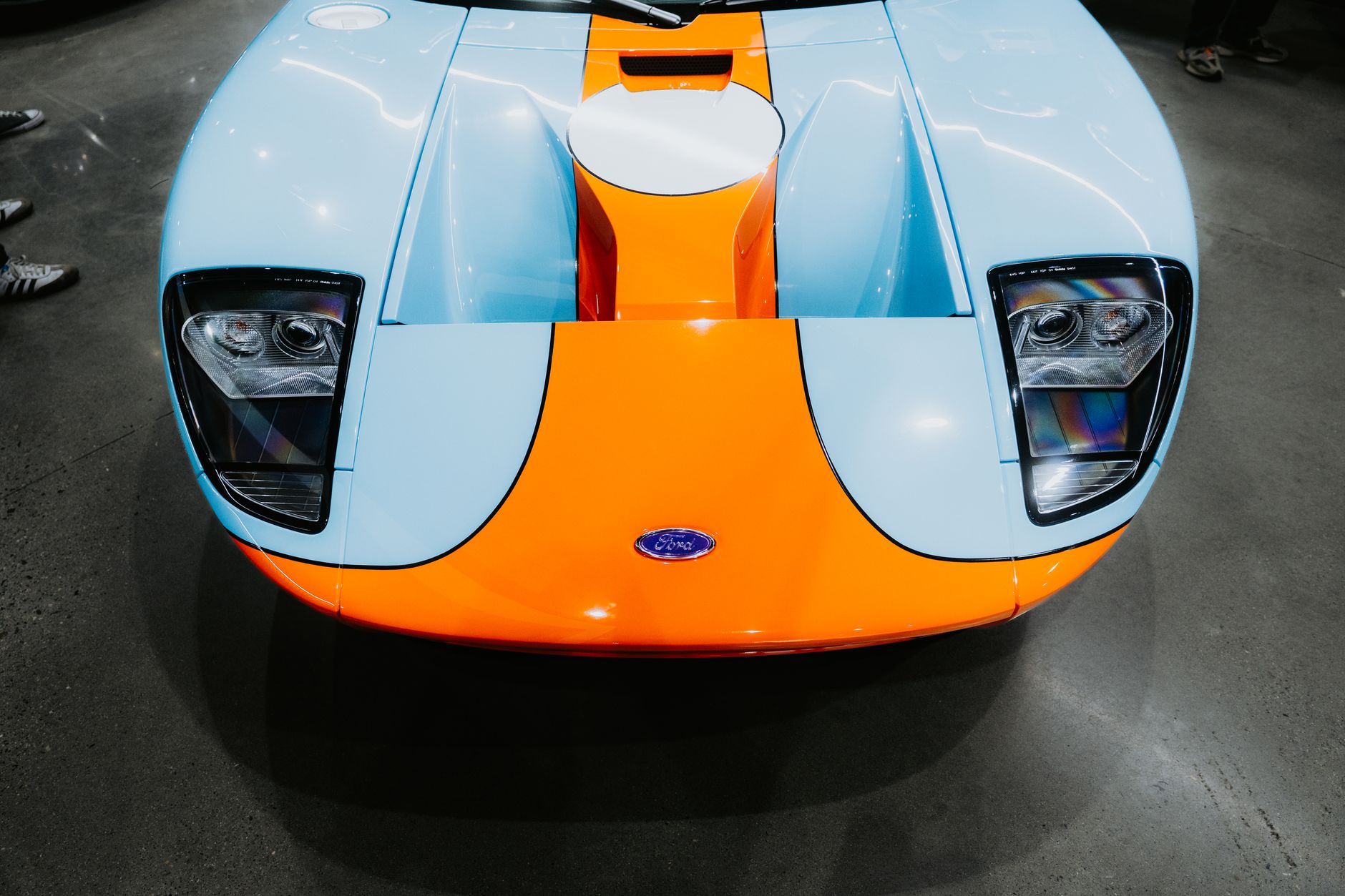 The front end of a low, wide two-seat sports car in pale blue with a broad orange centre stripe, photographed from above on a polished concrete floor
