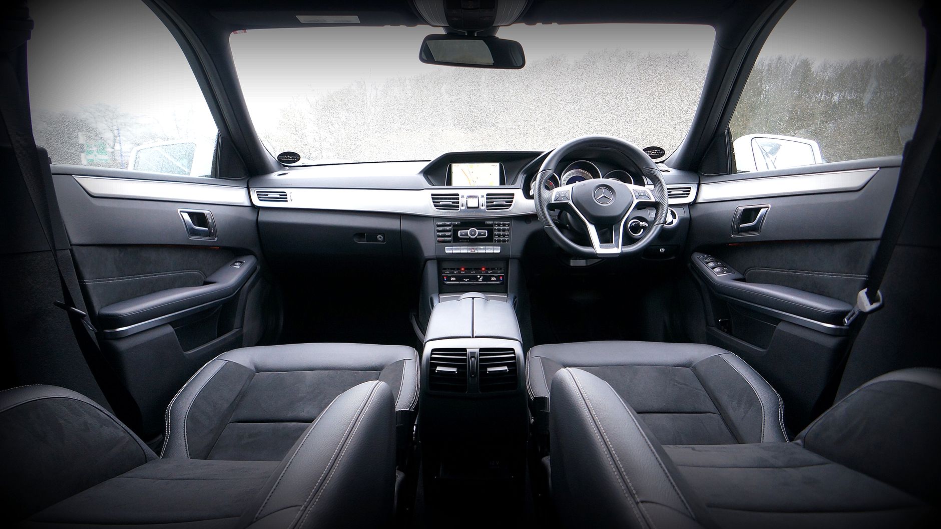 The cabin of a modern saloon seen from the back seat: black leather and suede seats with pale stitching, brushed metal trim across the dashboard and a navigation screen lit above the centre console