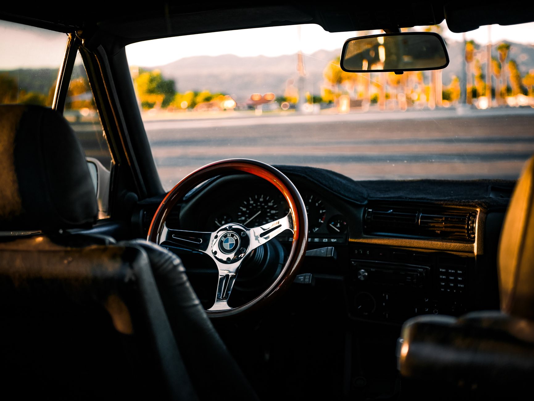 The interior of a classic coupe at golden hour, its wood-rimmed steering wheel in the foreground and palms and hills beyond the windscreen