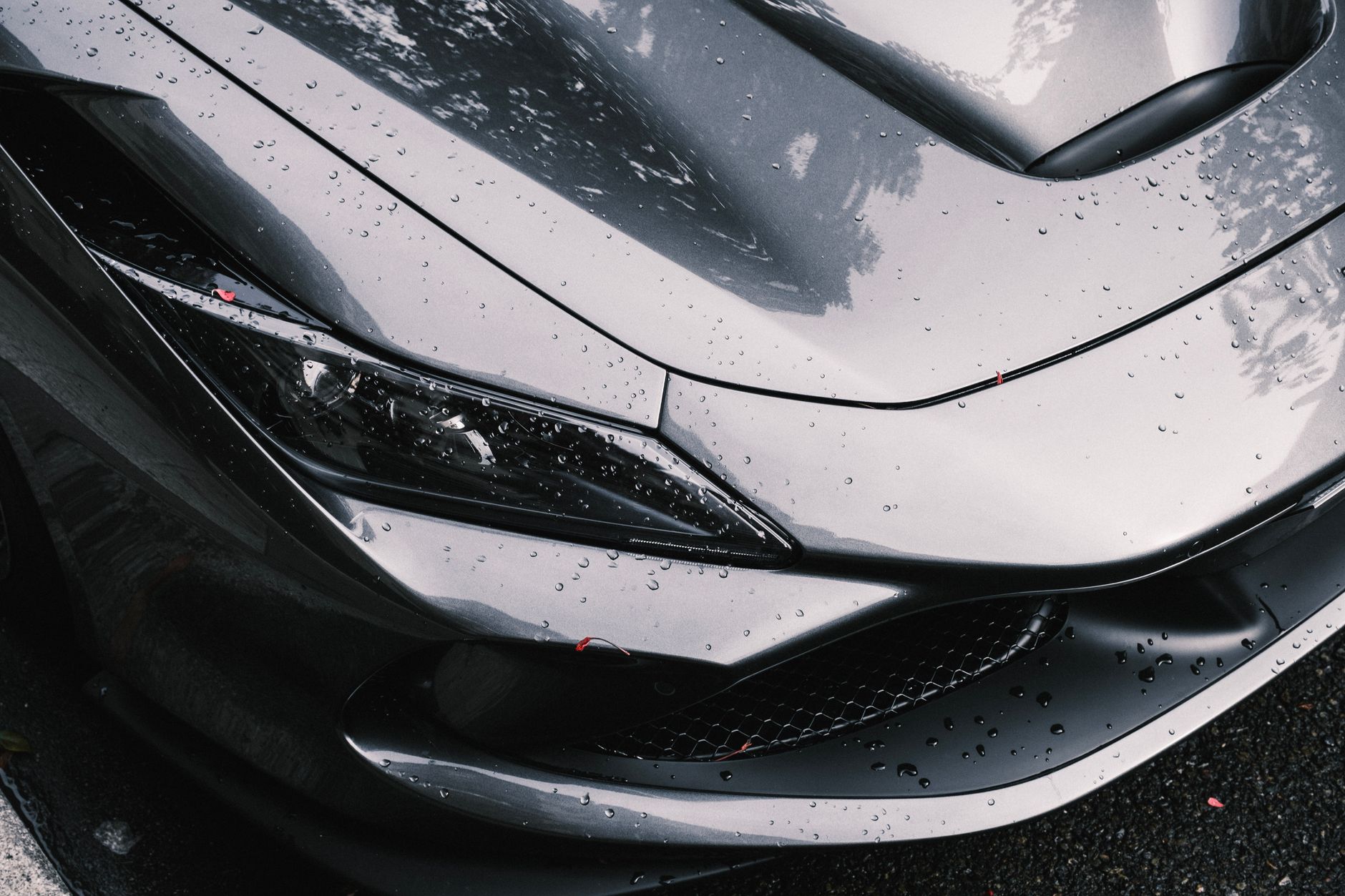 A close crop of the front of a dark grey sports car covered in rain droplets, showing the headlight, the bonnet vent and the front air intake