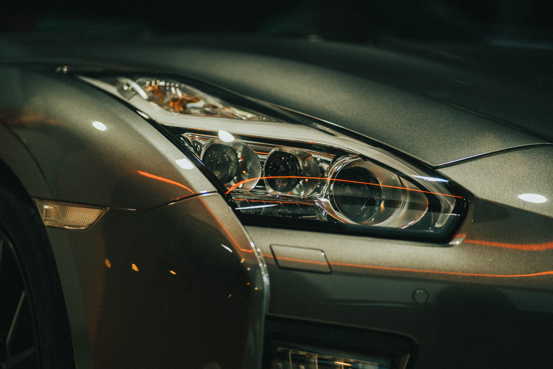 A close-up of the headlight and front wing of a dark grey sports car, lit by warm reflections at night