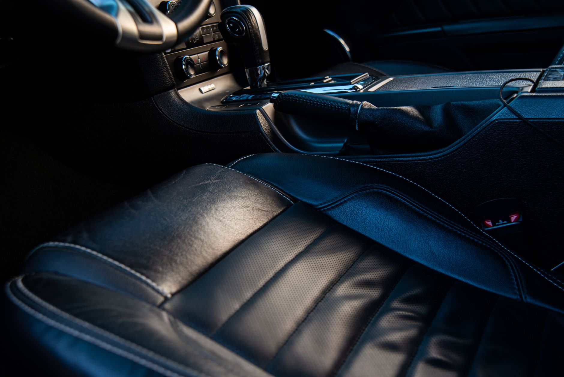 The interior of a car in low light, showing a stitched black leather driver's seat, the handbrake lever and part of the centre console and steering wheel