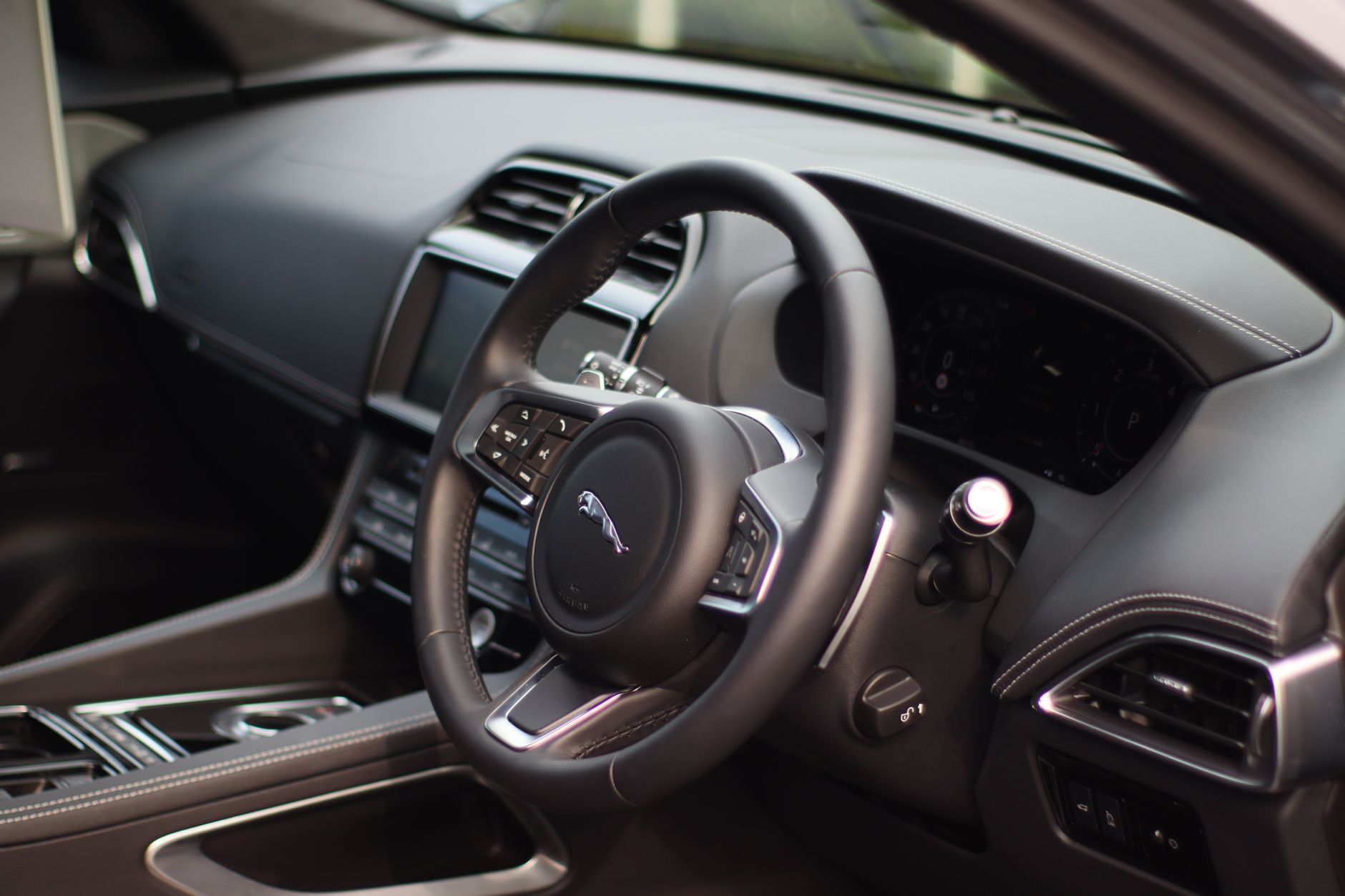 The interior of a right-hand-drive car, with a leather steering wheel, dark dashboard and centre console screen seen through the open driver's door