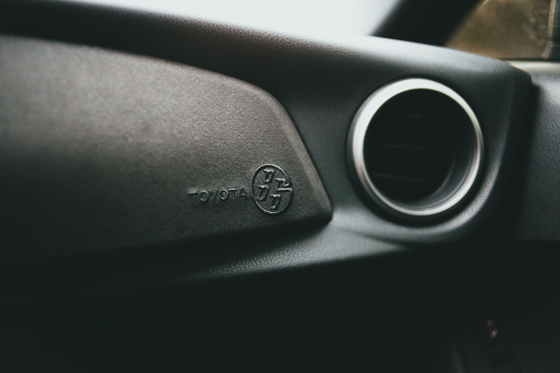 A softly lit close-up inside a car: a dark textured dashboard panel carrying an embossed manufacturer wordmark and roundel, a circular air vent ringed in chrome to the right, and a blurred strip of bright window along the top