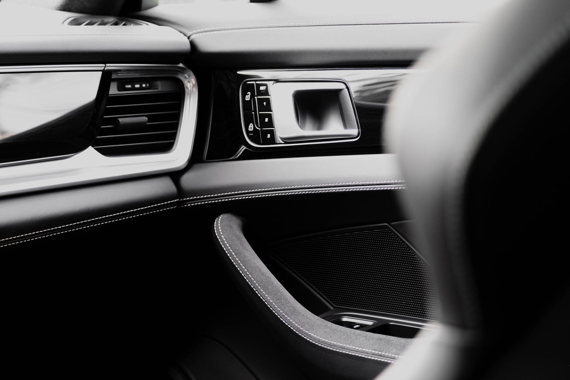 A monochrome close-up inside a car: the passenger door panel with stitched leather, a chrome-edged air vent, memory-seat buttons beside a recessed door handle, and a suede armrest, with the steering wheel rim blurred across the foreground