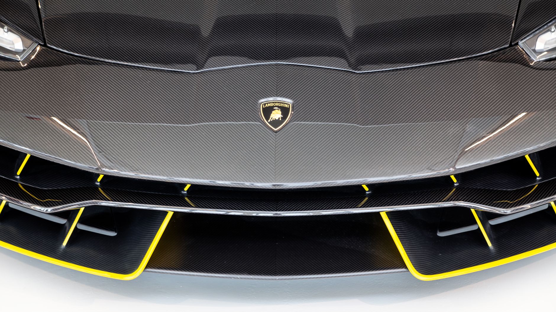 A close crop of the front of a black carbon-fibre supercar against a white studio wall, yellow trim running along the edges of the front splitter and air intakes