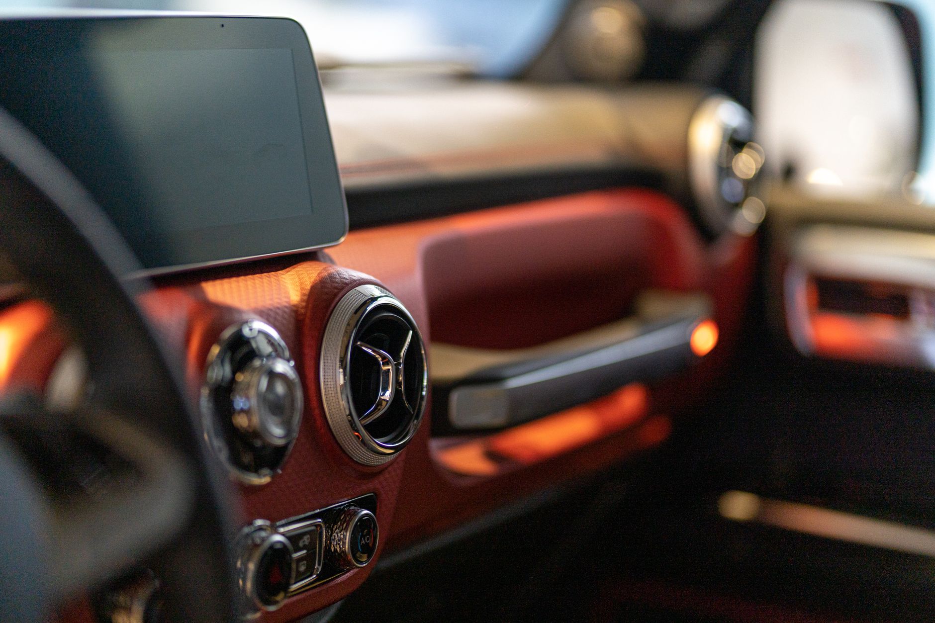 A close view of a car interior in a showroom, red leather across the dashboard and door, a chrome-ringed air vent, a dark centre screen and the edge of the steering wheel in the foreground