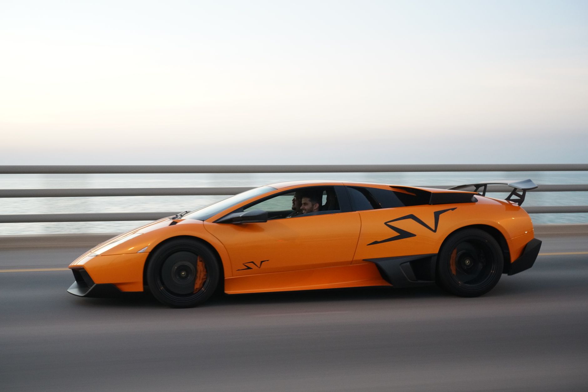 An orange sports car on a coastal road at dusk, with the sea and the last of the light behind it