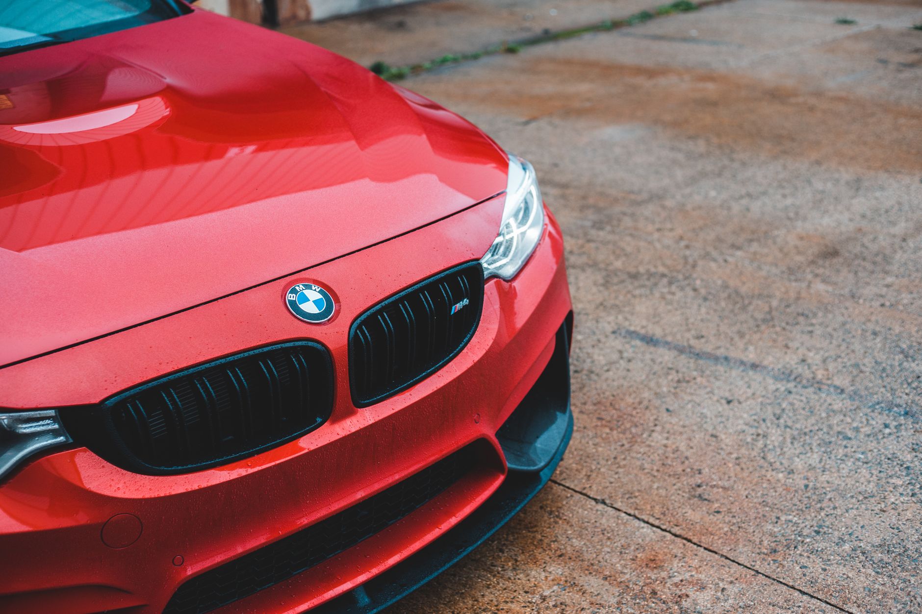 Close-up of the bonnet, black grille and headlight of a red performance coupé parked on a concrete forecourt, with rain spots still on the paintwork