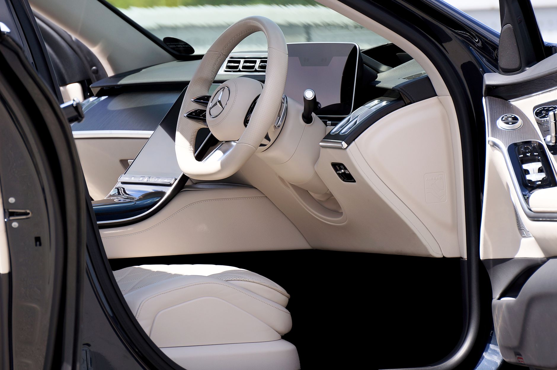 The open driver's door of a dark saloon car showing a cream leather interior, a perforated steering wheel and a tall touchscreen set into the dashboard