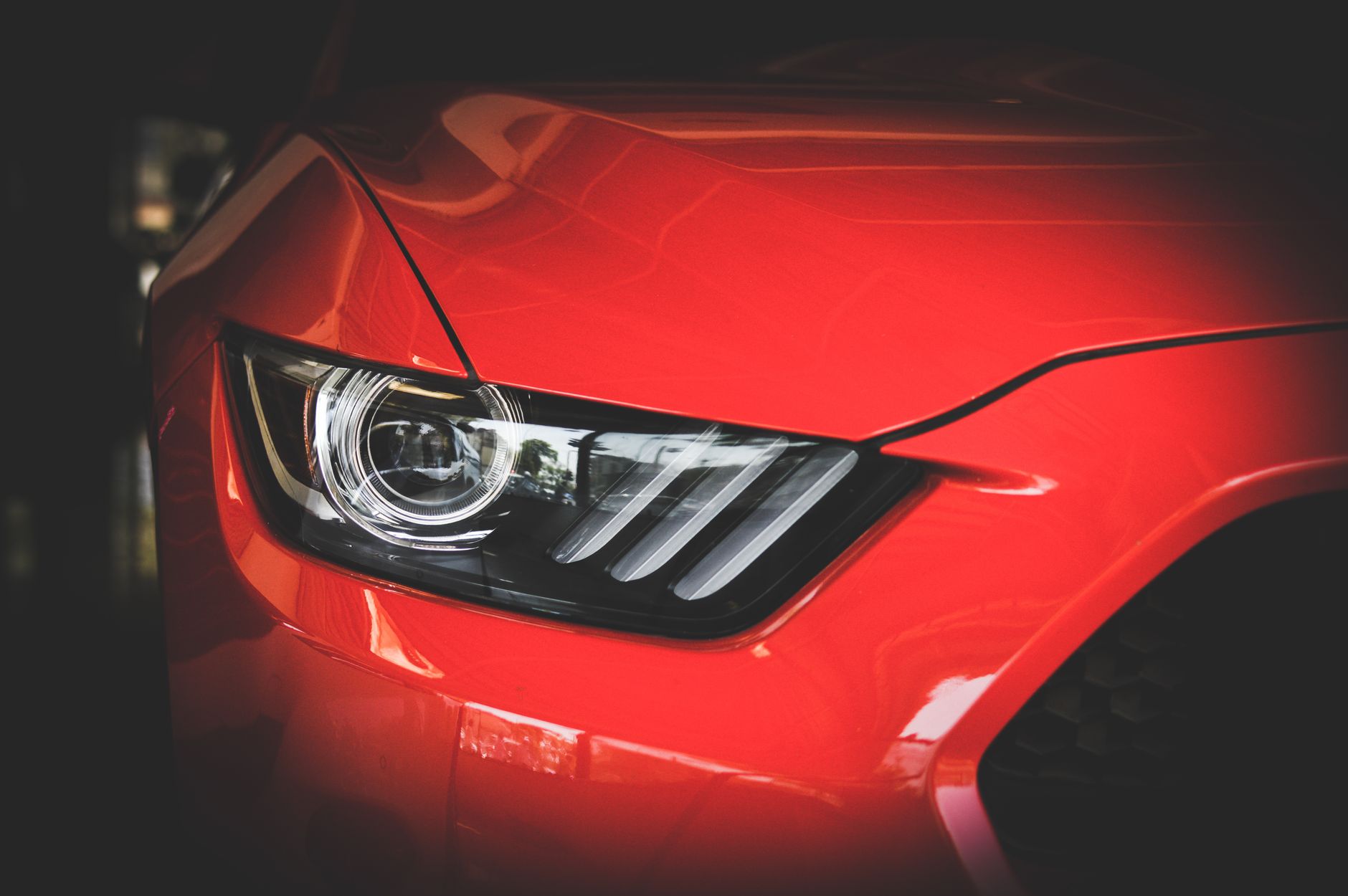 A close crop of the front corner of a bright red sports car in shadow, showing the headlight cluster and the sculpted line of the wing