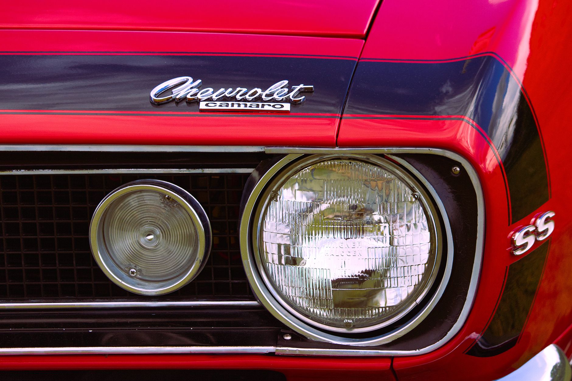 A tight close-up of the front corner of a bright red classic muscle car, showing the chrome-ringed round headlamp, the mesh grille and the polished script badge on the fender