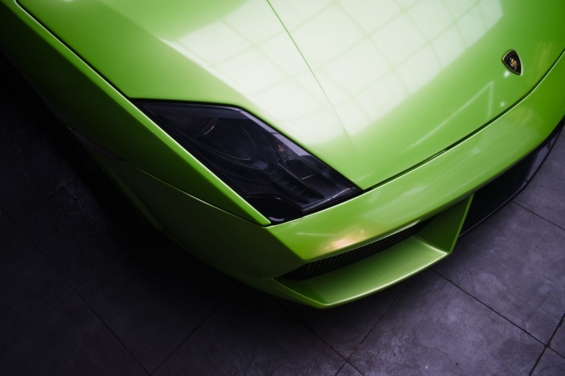 Close crop of the front of a lime-green sports car on a dark tiled floor, showing the angular headlight, the sculpted bonnet line and the low front splitter