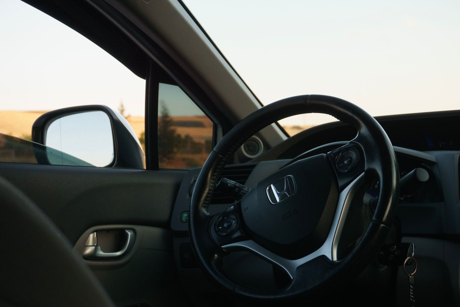 The interior of a car at golden hour, looking past the steering wheel and wing mirror to dry summer fields and a pale sky