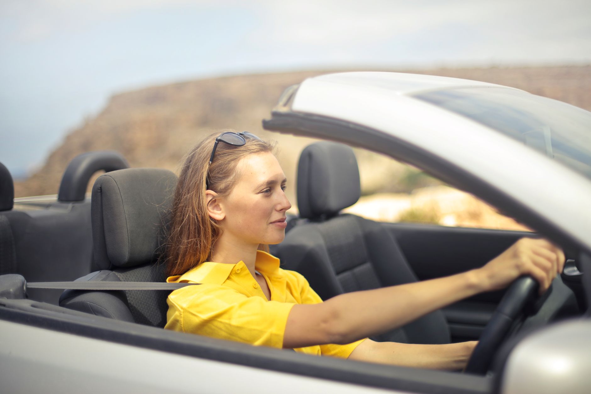 A woman in a yellow shirt driving a silver convertible with the roof down, a dry coastal headland blurred behind her