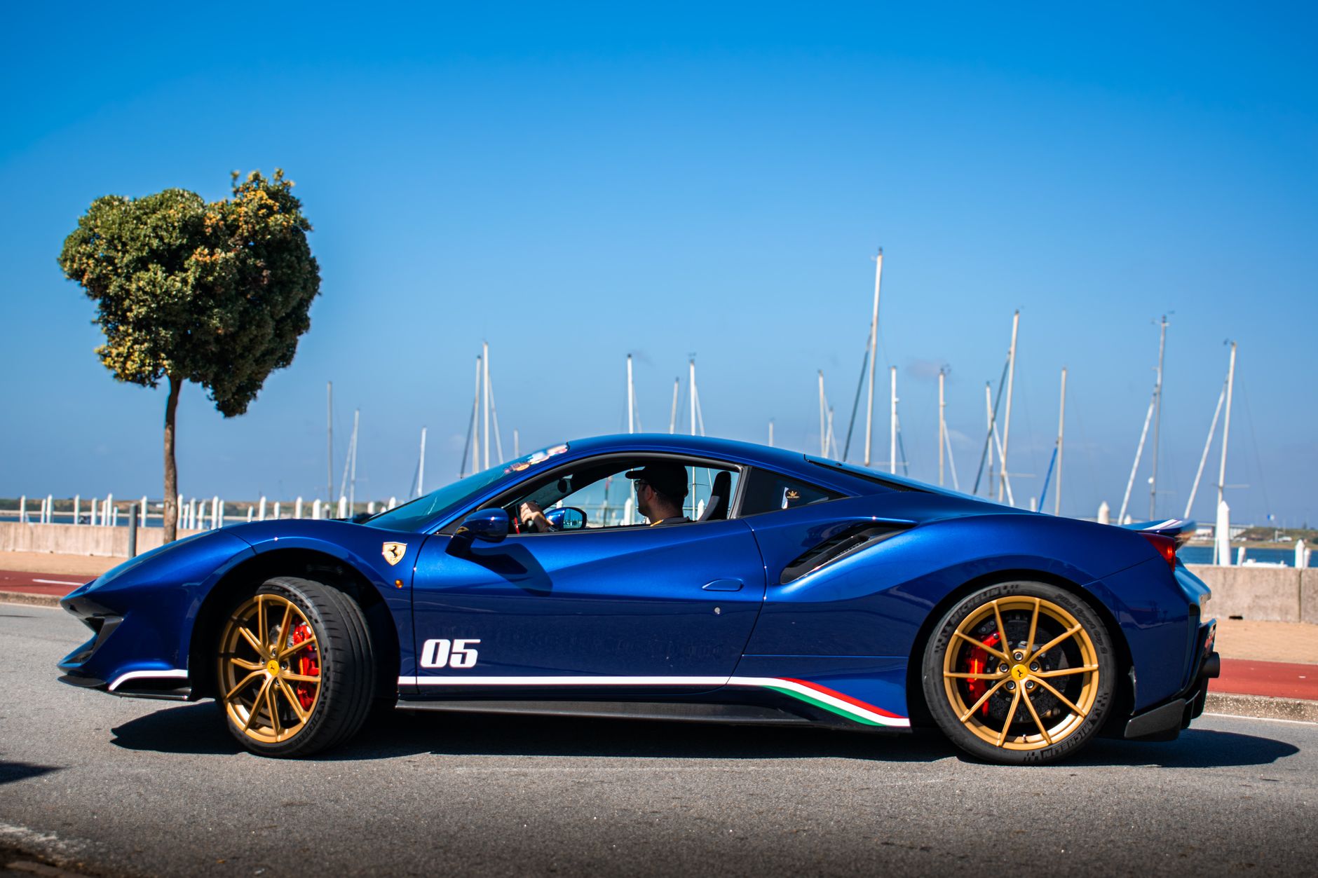 A blue sports car with gold wheels driving past a marina full of moored sailing yachts under a clear sky