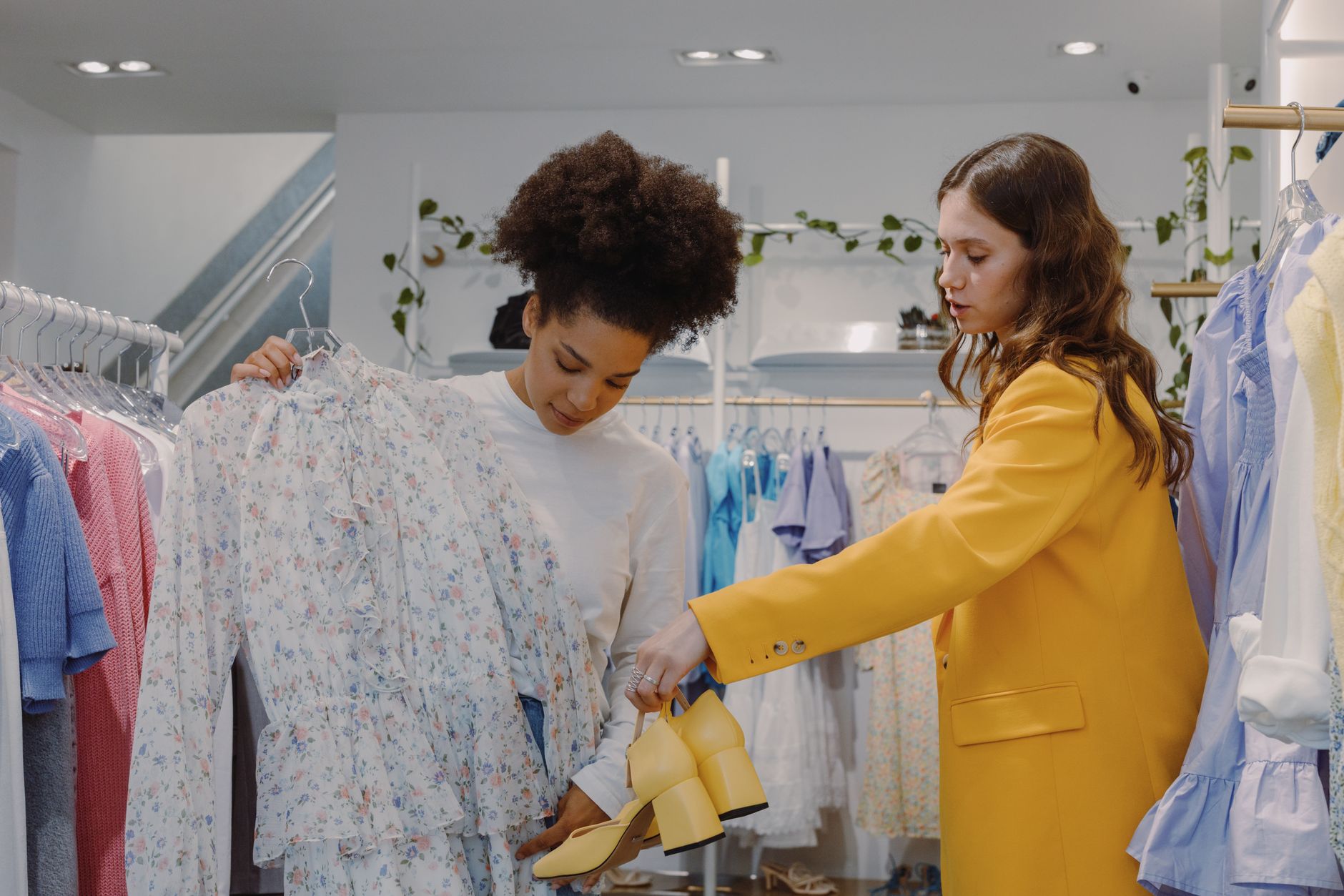 Two women in a bright clothing store, one holding a floral dress on a hanger while the other offers a pair of yellow heeled shoes