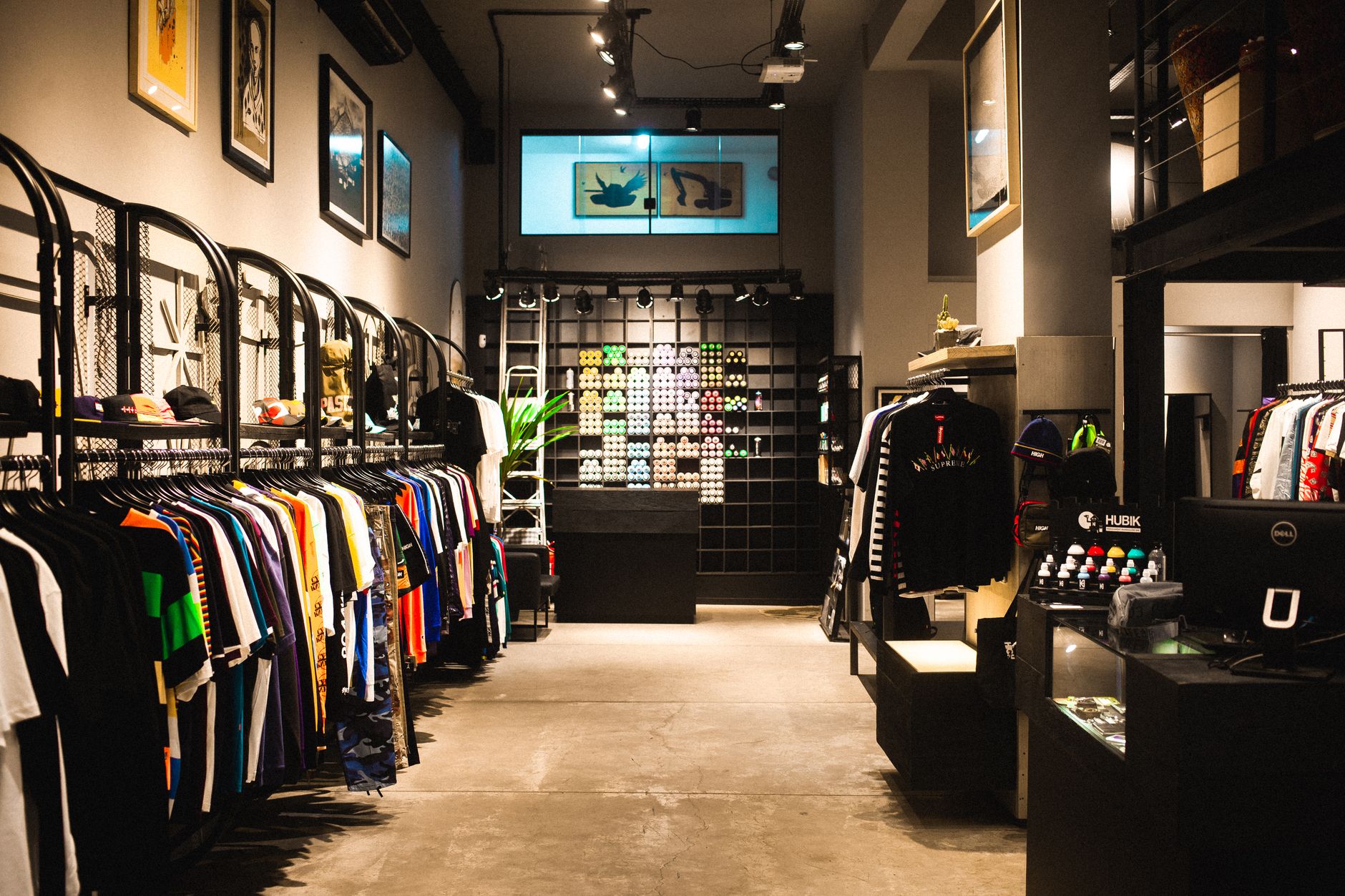 The interior of an independent clothing boutique: rails of brightly printed t-shirts, caps on a shelf, framed prints on grey walls and a backlit display wall under spotlights