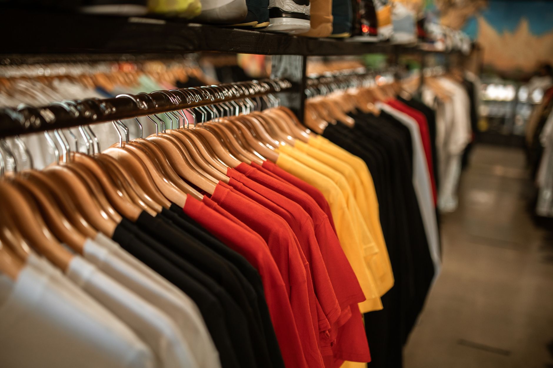 A long rail of t-shirts and polo shirts in white, black, red and yellow hanging on wooden hangers, with shoes on a shelf above and the rest of the shop out of focus behind