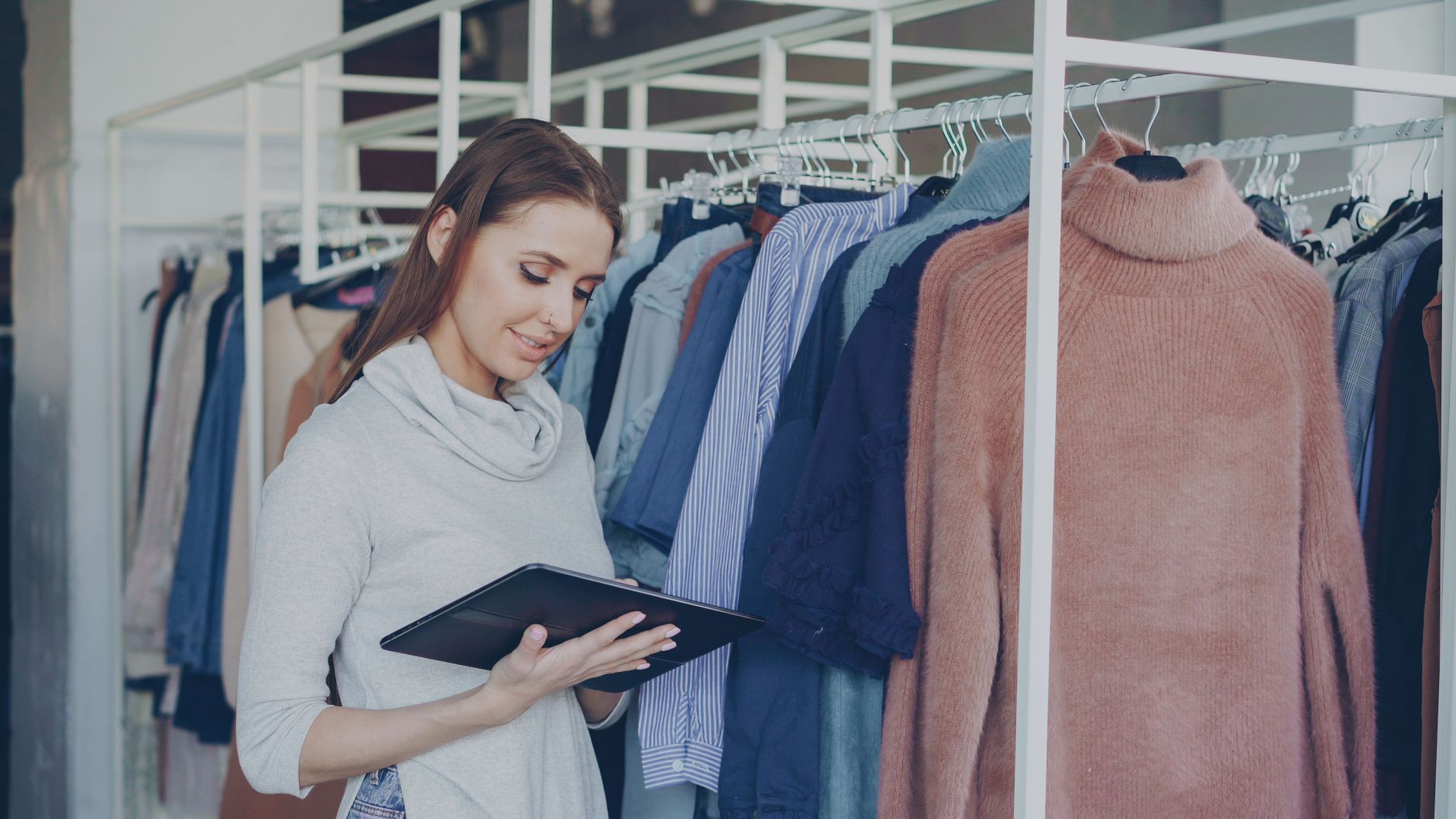 A woman in a light grey top holding a tablet while checking stock on a rail of clothes in a bright boutique