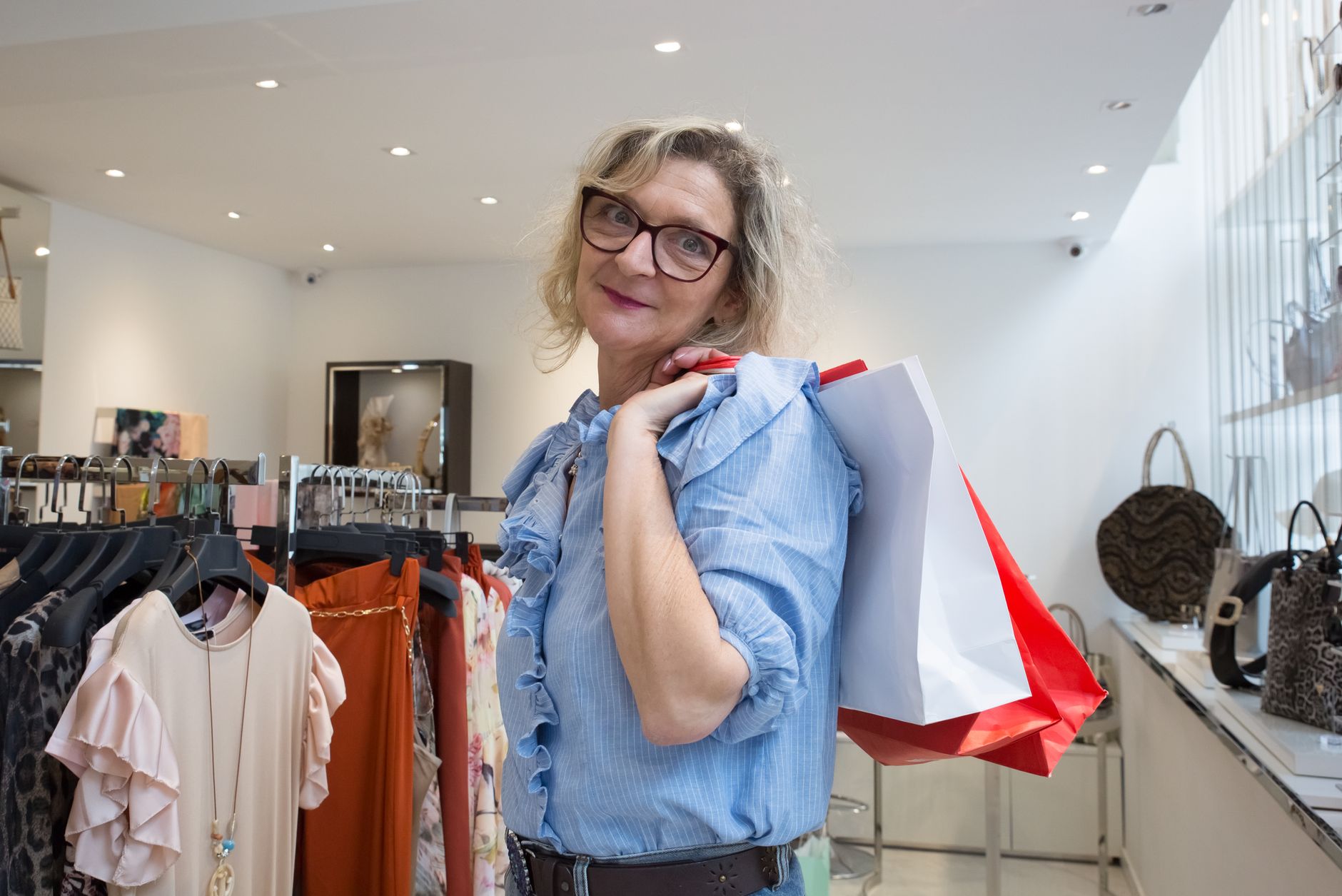 A woman in a blue ruffled blouse and dark-rimmed glasses carrying red and white paper bags over her shoulder in a bright white boutique, clothes rails and handbags on either side