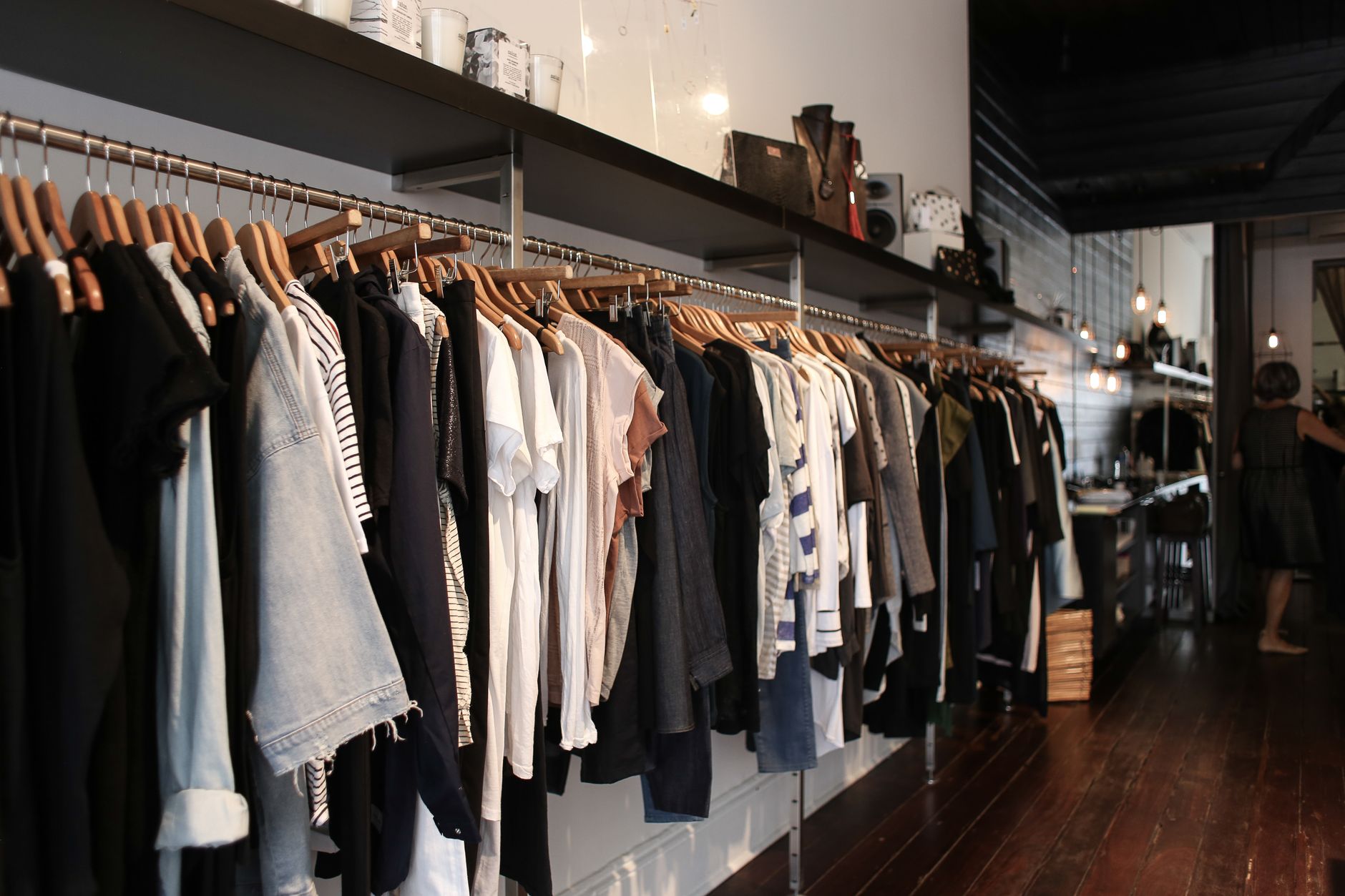 A long rail of shirts, jackets and knitwear on wooden hangers in a warmly lit independent boutique with dark timber floors and pendant lamps