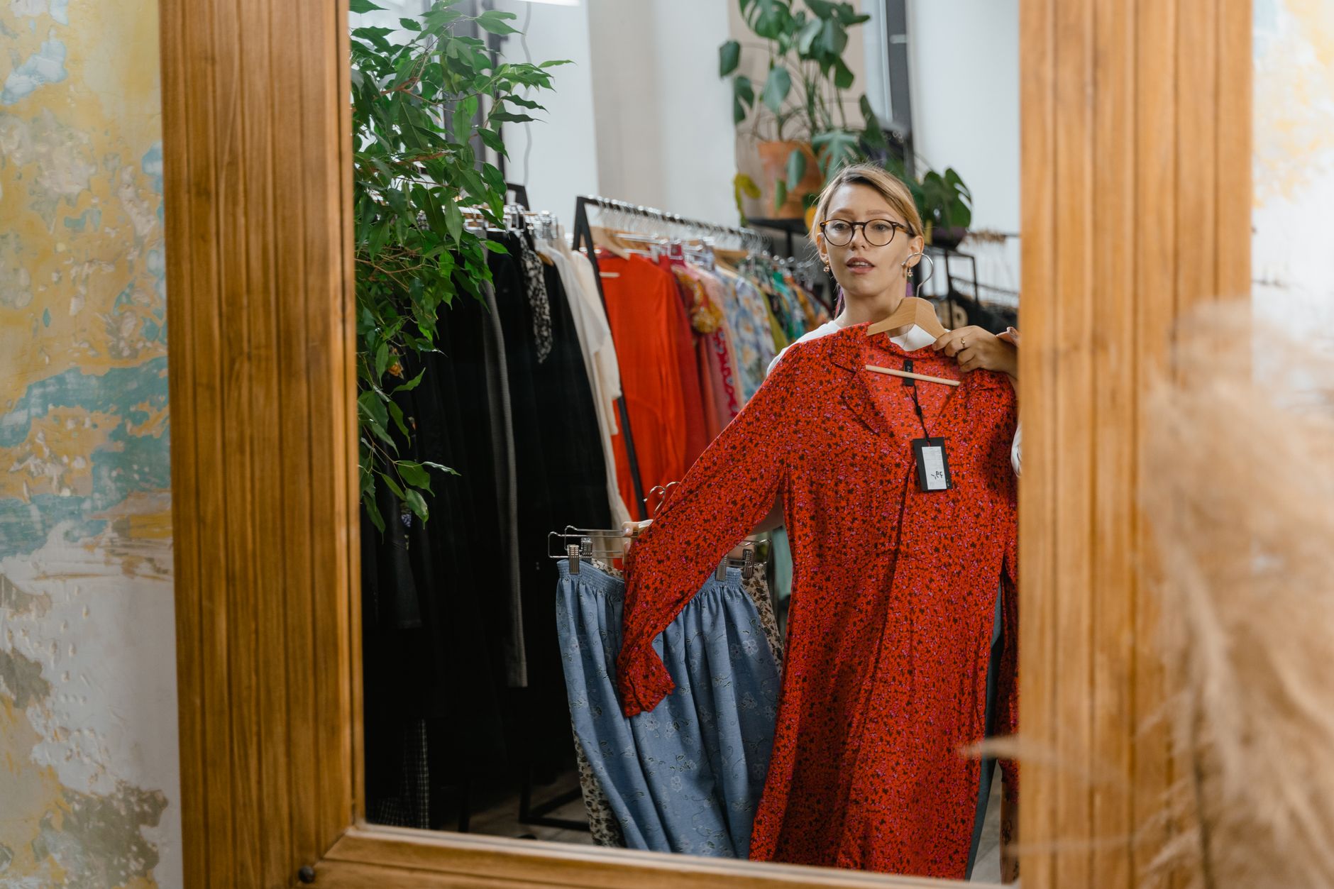A woman in glasses holding a red floral dress on a hanger up against herself in front of a wooden-framed mirror in a boutique