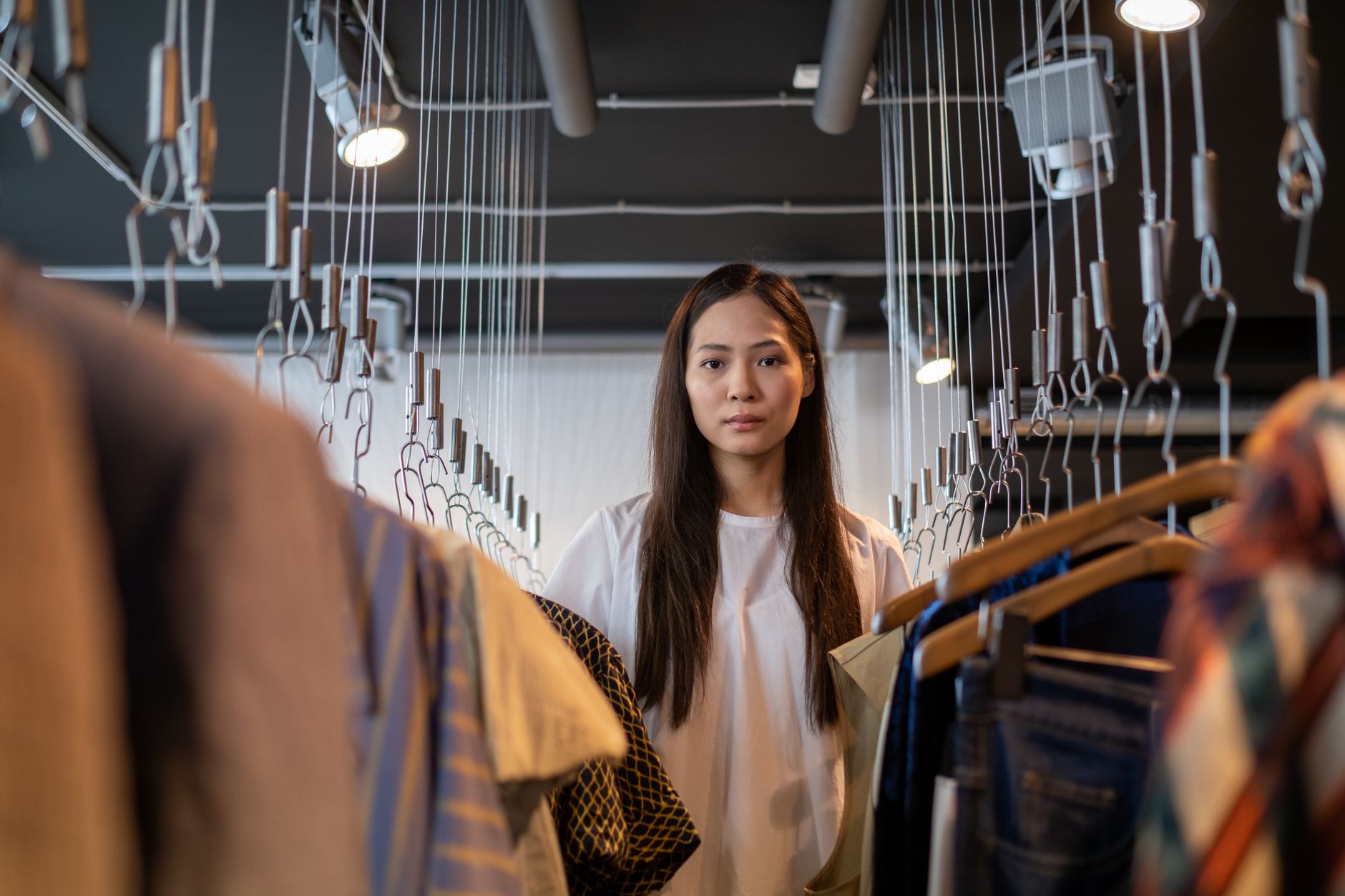 A woman standing between rails of clothes in a dark modern boutique lit by spotlights on suspended cables