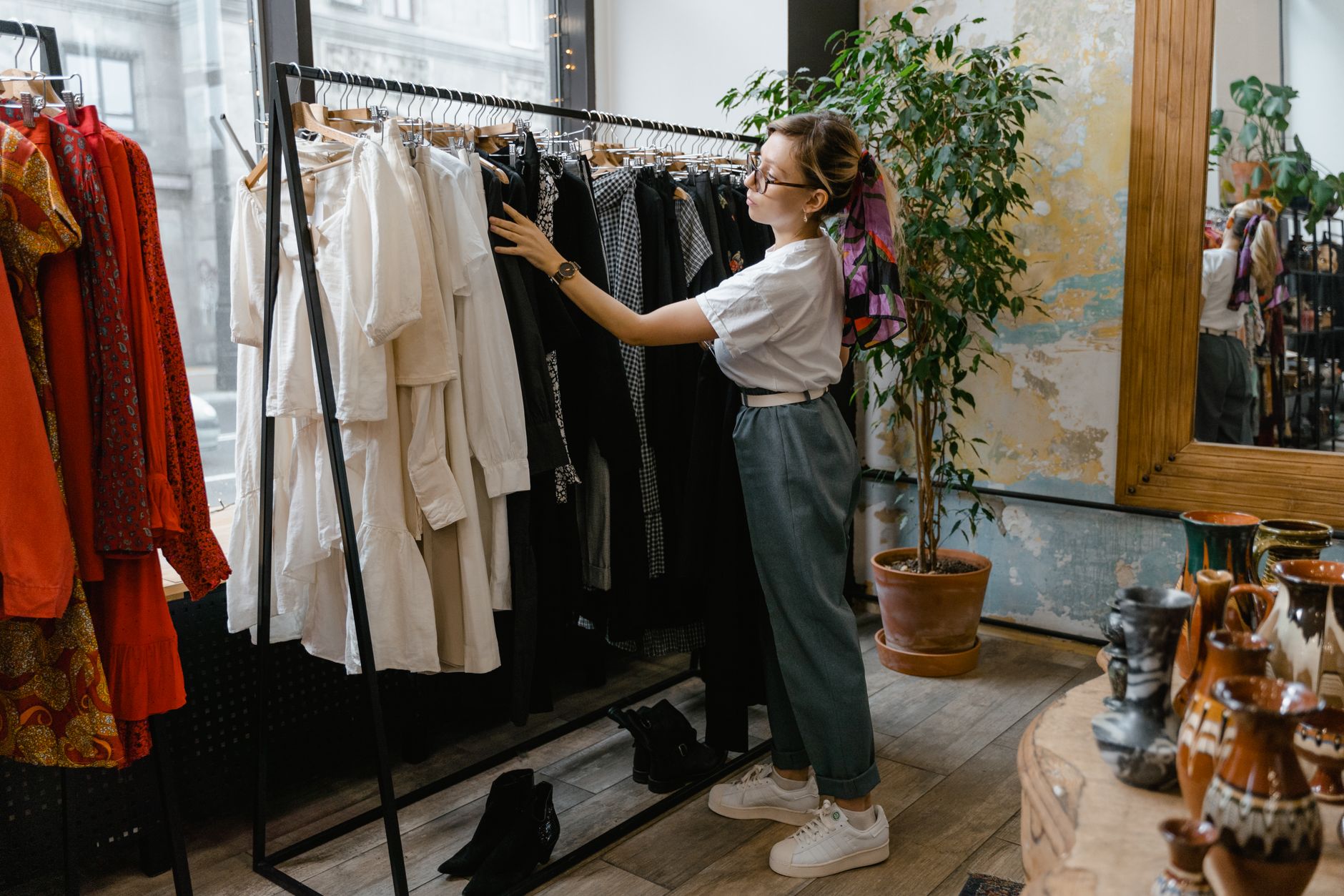 A woman looking through a rail of clothes in a bright boutique, with potted plants and ceramics on the shelves behind her